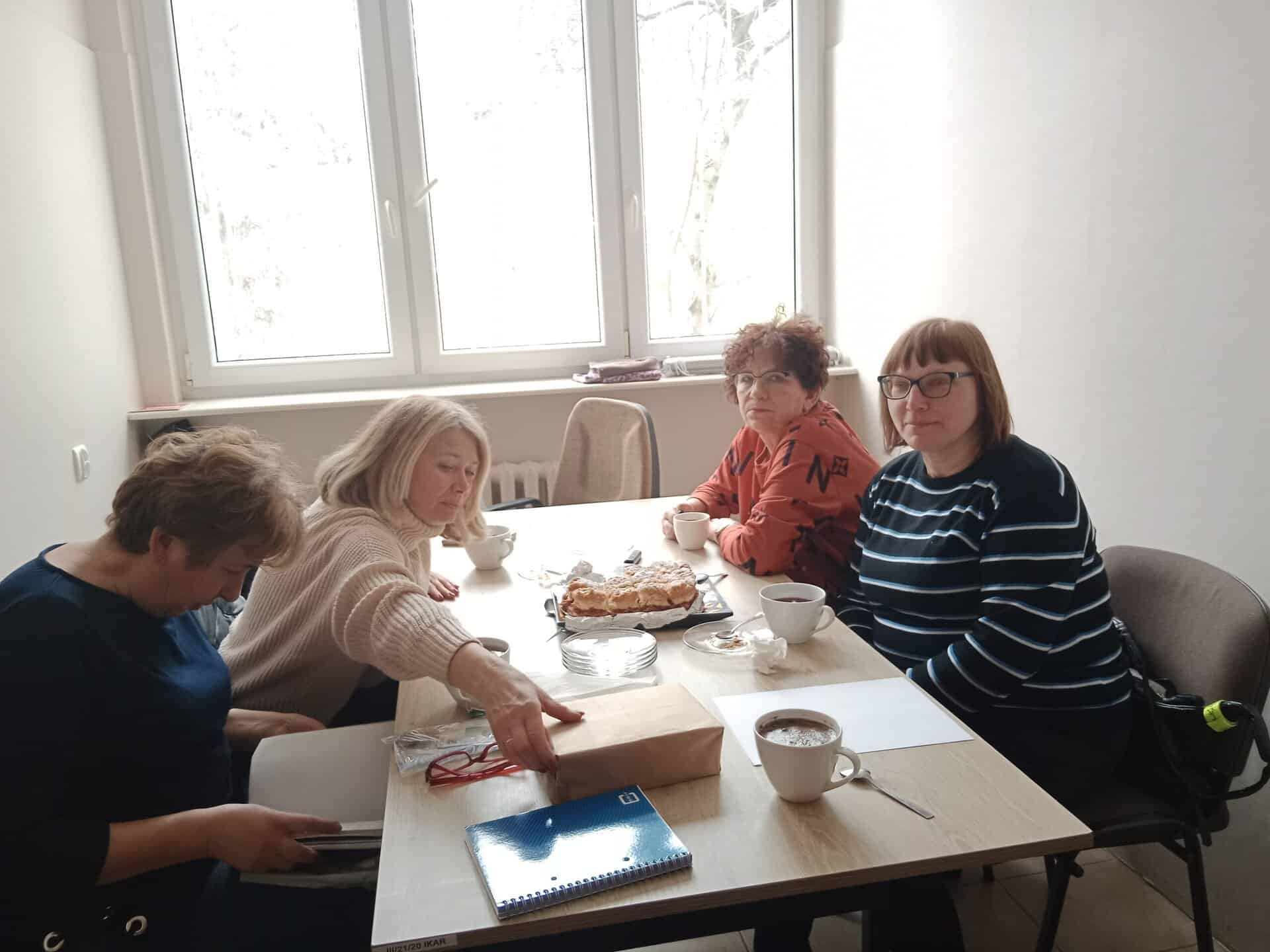 Cztery kobiety siedzą przy stole z kawą, gazetami i ciastem w jasno oświetlonym pokoju z dużym oknem.