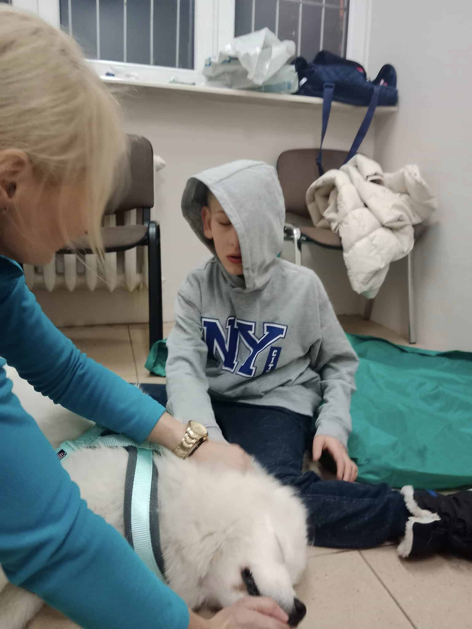 Dziecko w szarej bluzie NYY siedzi na podłodze obok kobiety głaszczącej puszystego białego psa w pomieszczeniu z krzesłami, płaszczami i zieloną matą.