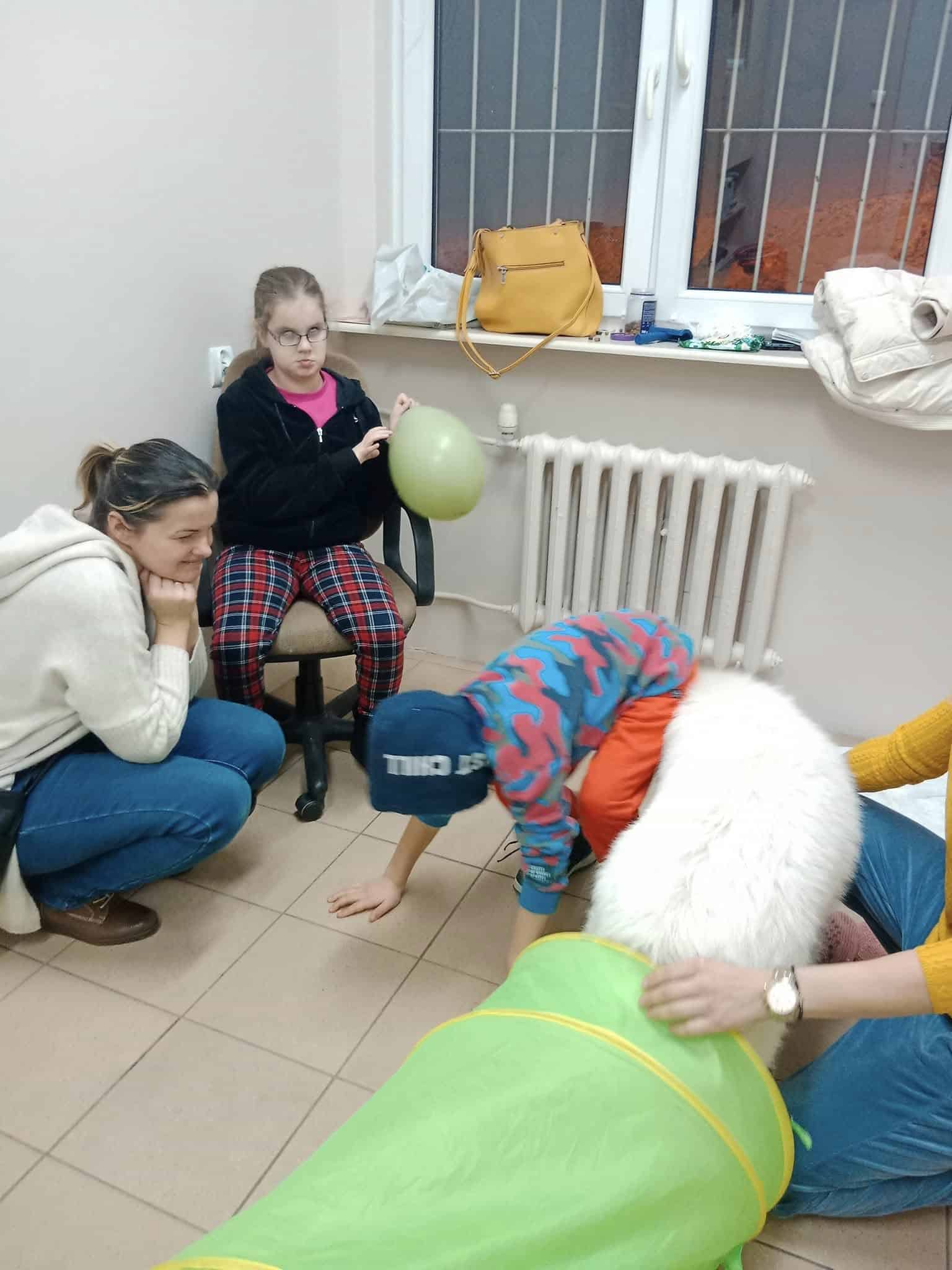 Cztery osoby, w tym dwoje dzieci i dwoje dorosłych, wchodzą w interakcję w pokoju; jedno dziecko trzyma balon, podczas gdy inne czołga się przez tunel do zabawy. Przedmioty spoczywają na grzejniku pod oknem.