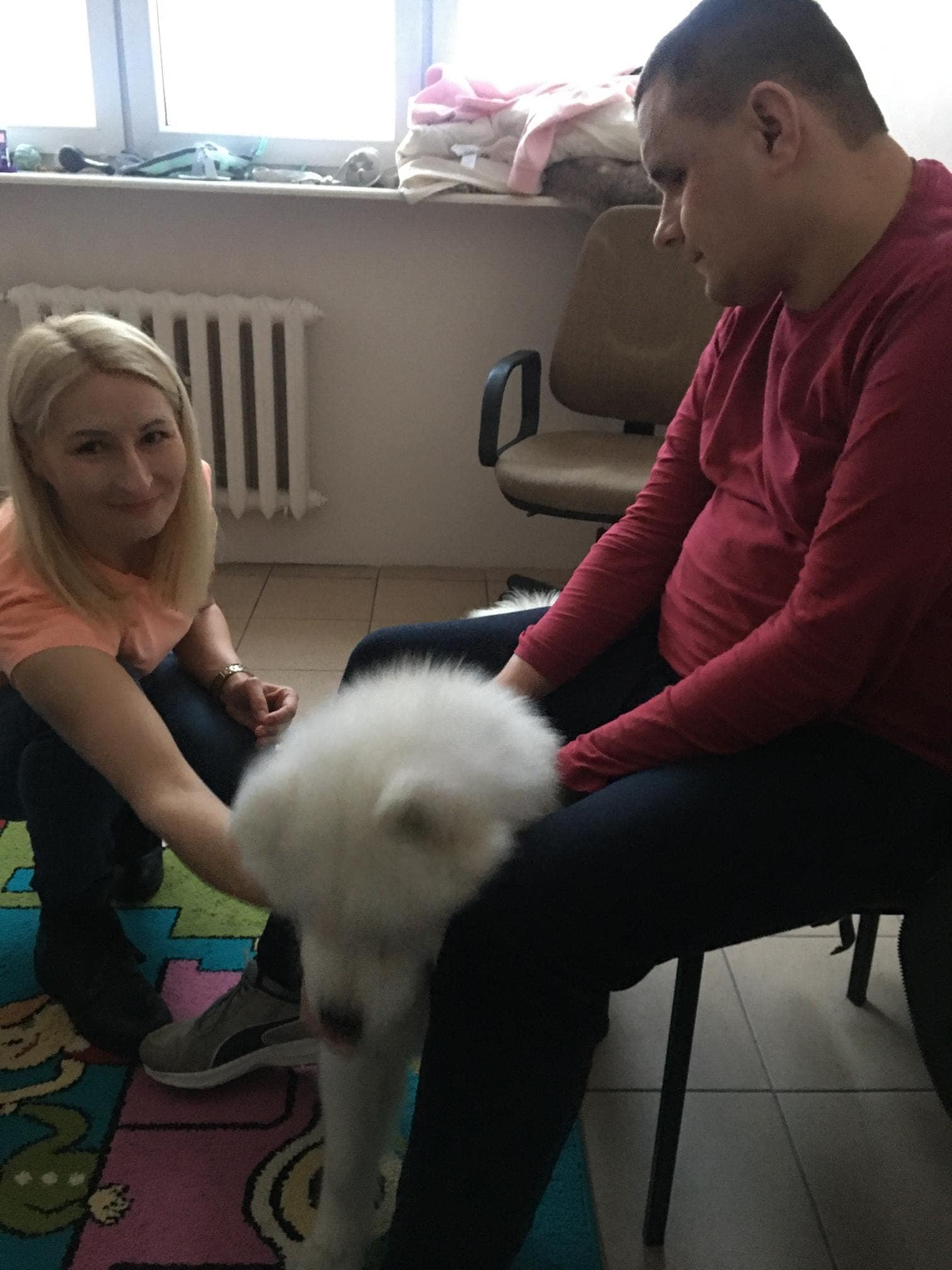 Kobieta klęczy na podłodze, a mężczyzna siedzi na krześle; oboje wchodzą w interakcję z puszystym białym psem na kolorowym dywanie w pokoju z podłogą wyłożoną kafelkami i kaloryferem.