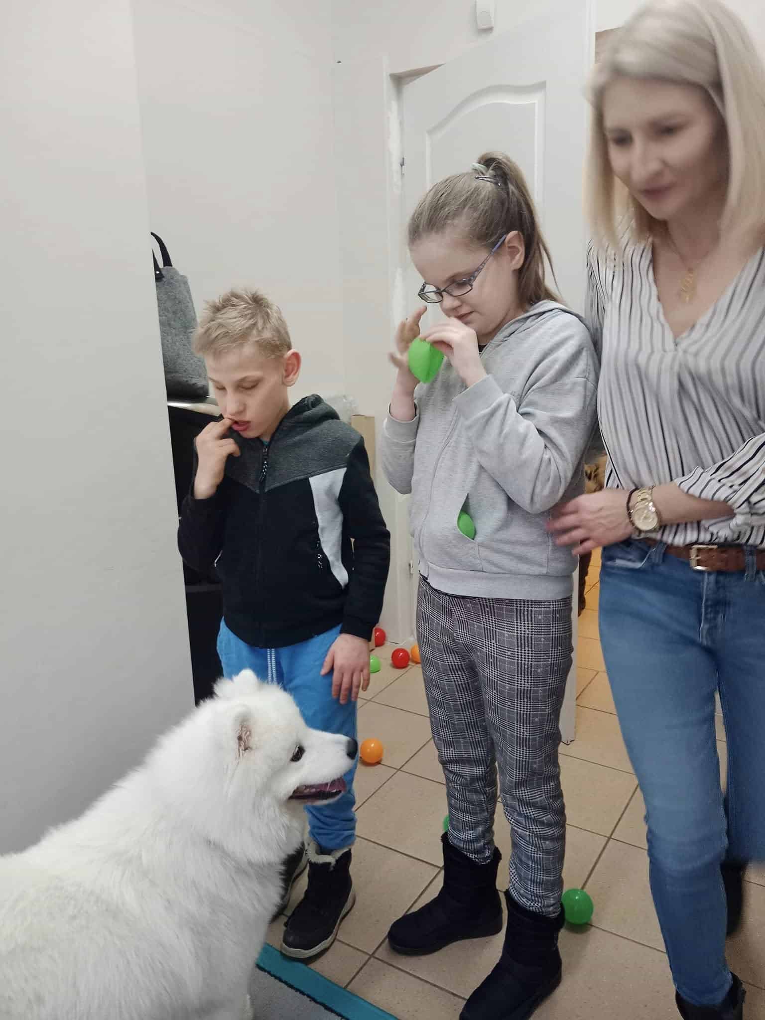 Kobieta stoi obok dwójki dzieci, z których jedno trzyma zielony balonik, podczas interakcji z białym puszystym psem w pomieszczeniu. Kolorowe piłki są rozrzucone na podłodze.