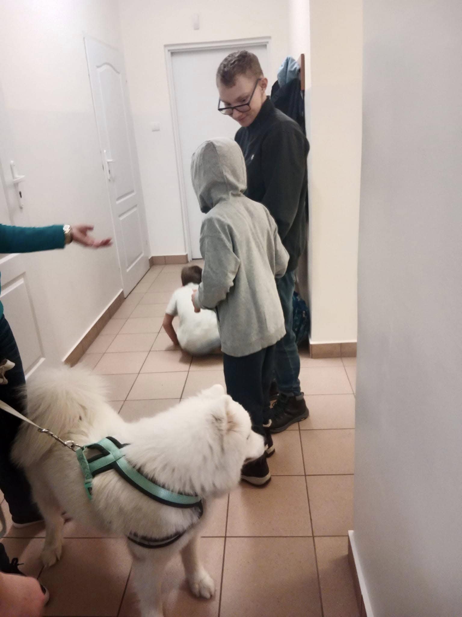 Biały pies na smyczy stoi w korytarzu z czterema osobami; jedna osoba siedzi na podłodze, a trzy inne stoją w pobliżu.