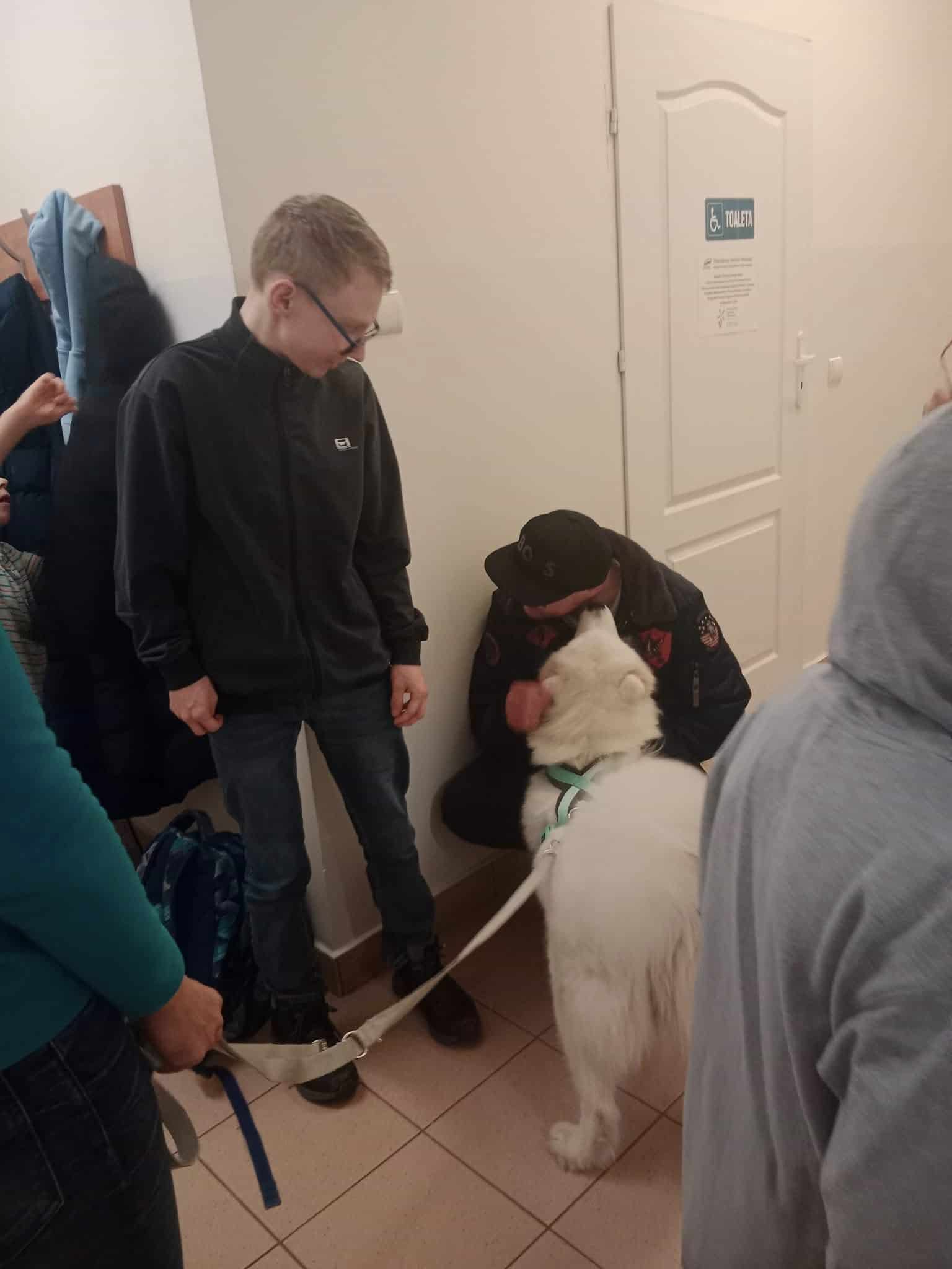 Kilka osób zebrało się w korytarzu; jedna osoba głaszcze dużego białego psa na smyczy, podczas gdy inna stoi w pobliżu, trzymając smycz.