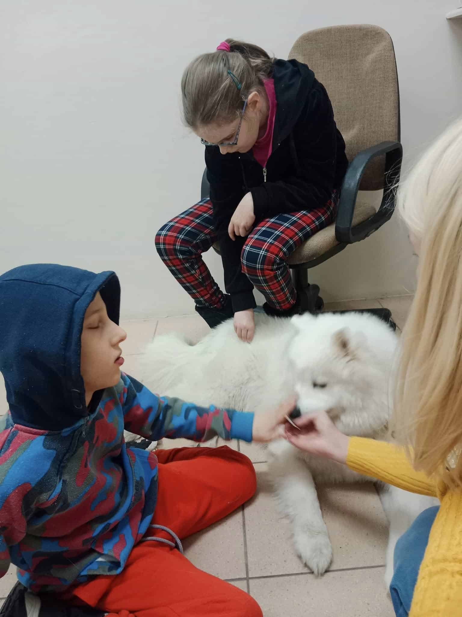 Chłopiec i dziewczynka wchodzą w interakcję z puszystym białym psem na podłodze, podczas gdy dorosły w żółtym swetrze nadzoruje. Dziewczynka siedzi na krześle, a chłopiec na podłodze.