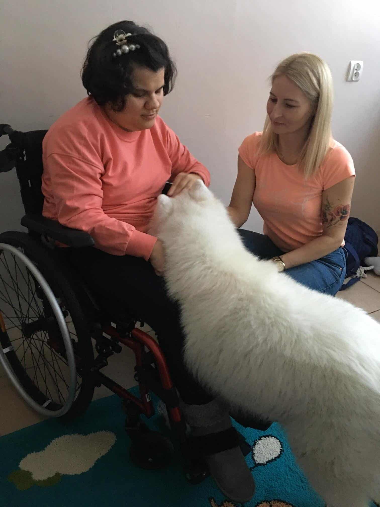 Kobieta na wózku inwalidzkim głaszcze dużego, puszystego białego psa, podczas gdy inna kobieta siedzi obok, uśmiechając się.