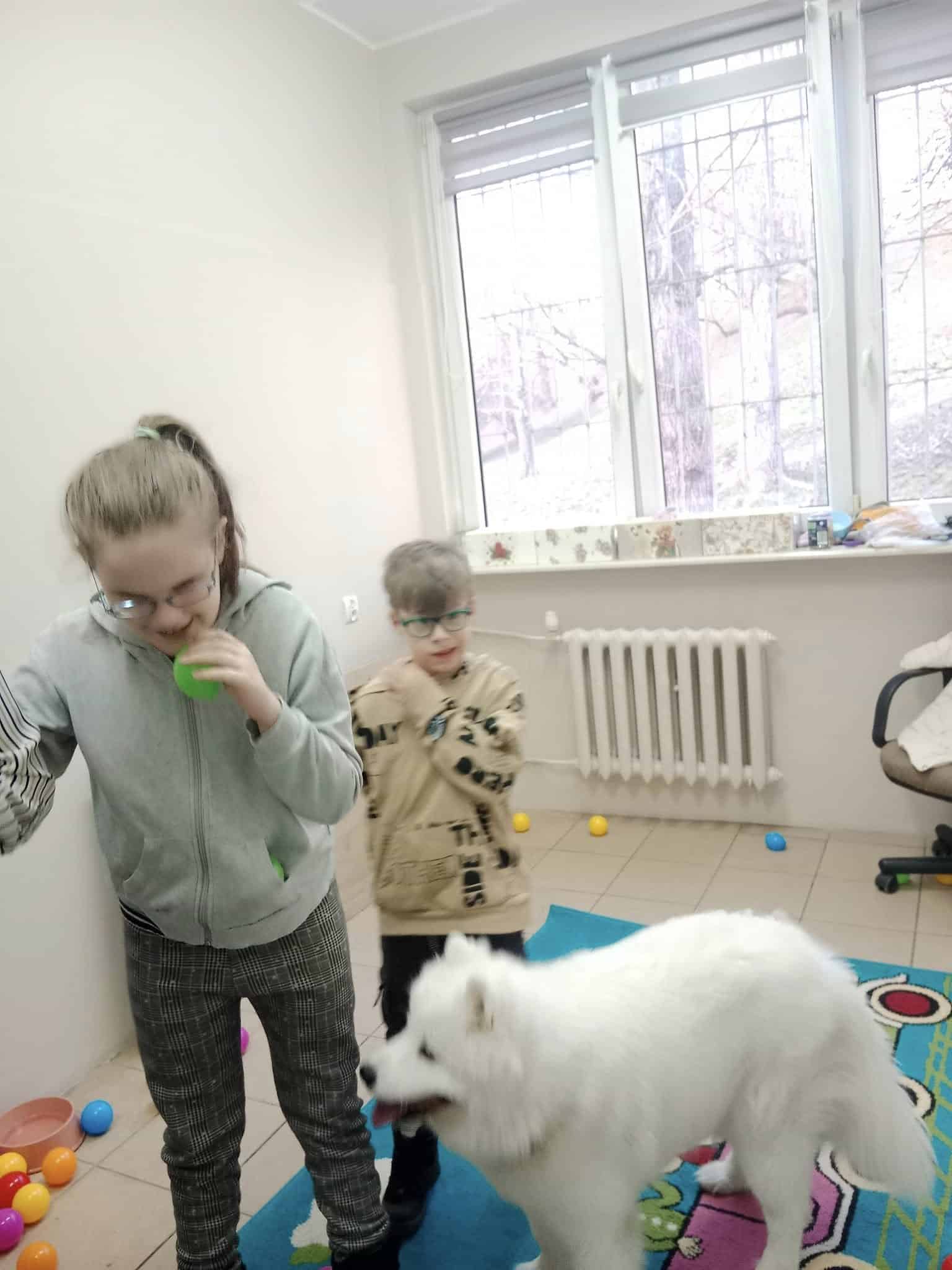 Dwoje dzieci i biały puszysty pies znajdują się w pokoju zabaw z kolorowymi plastikowymi piłkami rozrzuconymi na podłodze.