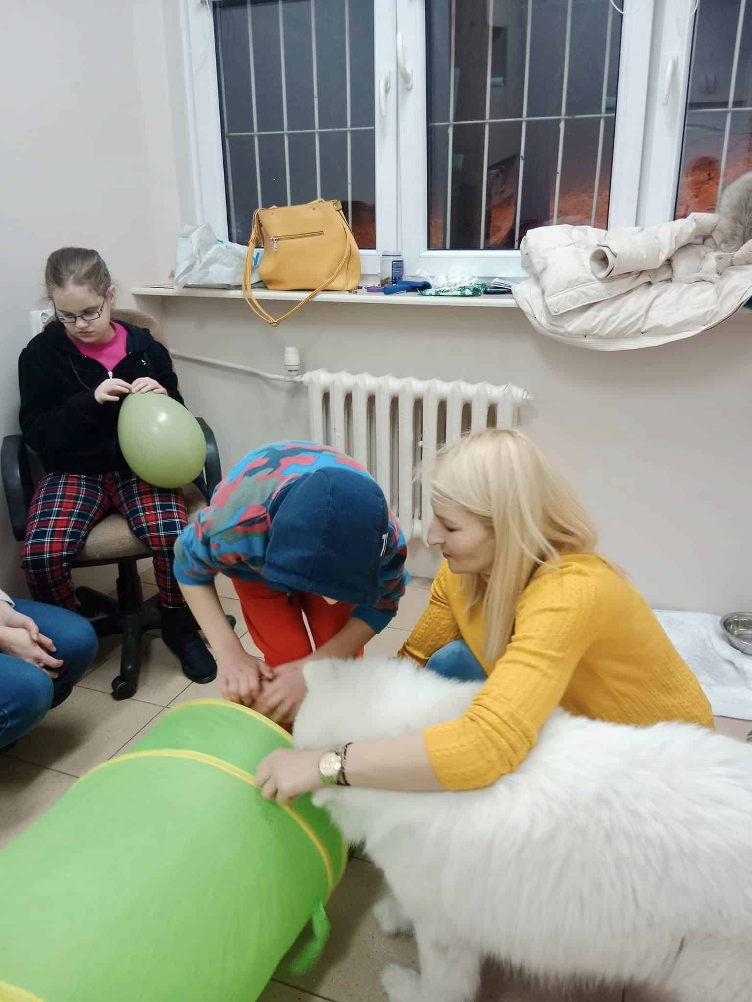 Kobieta w żółtym swetrze wchodzi w interakcję z dużym białym psem w pobliżu zielonego tunelu, podczas gdy dwoje dzieci siedzi w pobliżu, jedno trzyma zielony balon.