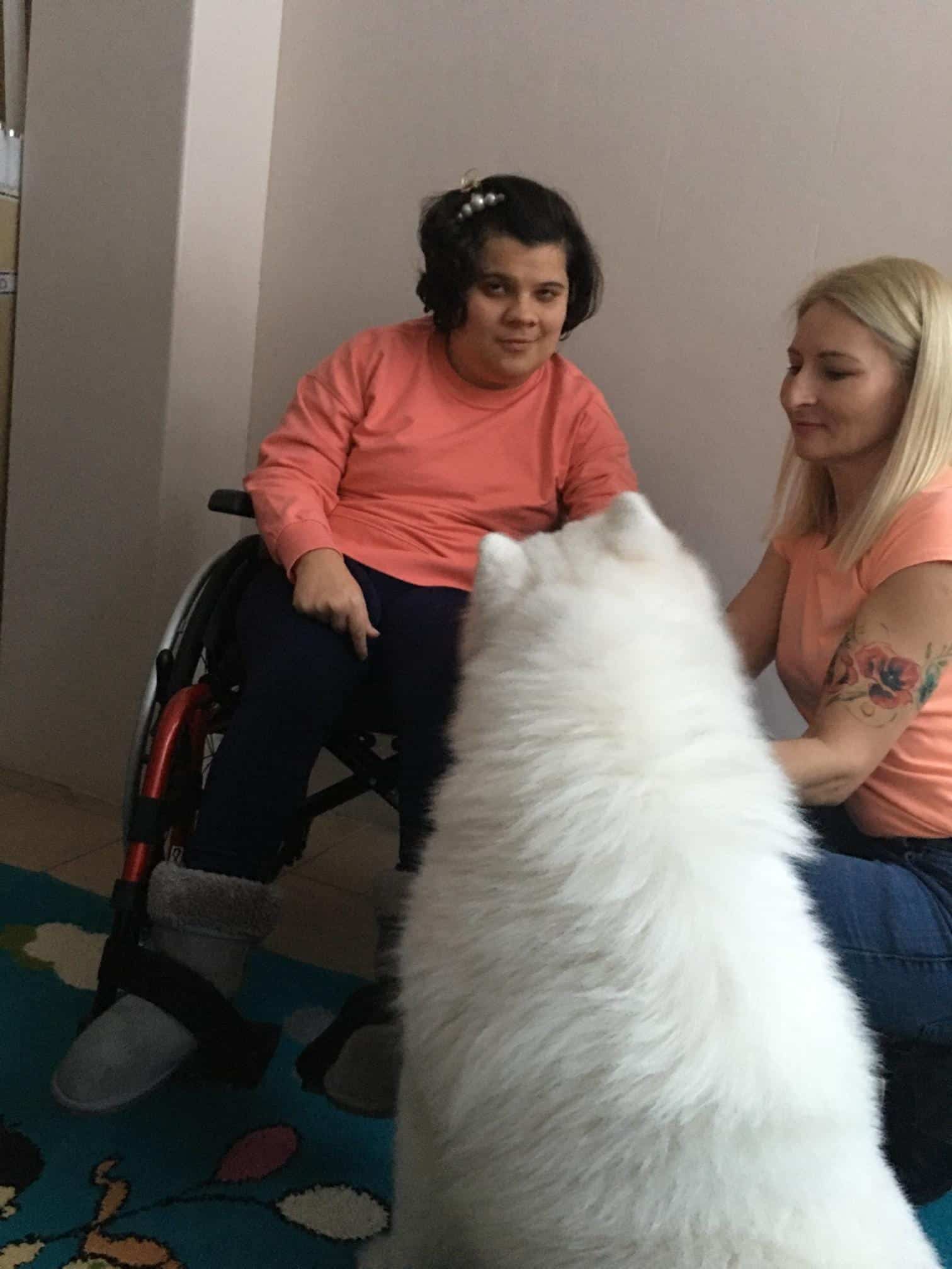 Dziewczyna na wózku inwalidzkim i kobieta siedzą na dywanie naprzeciwko dużego, puszystego białego psa.