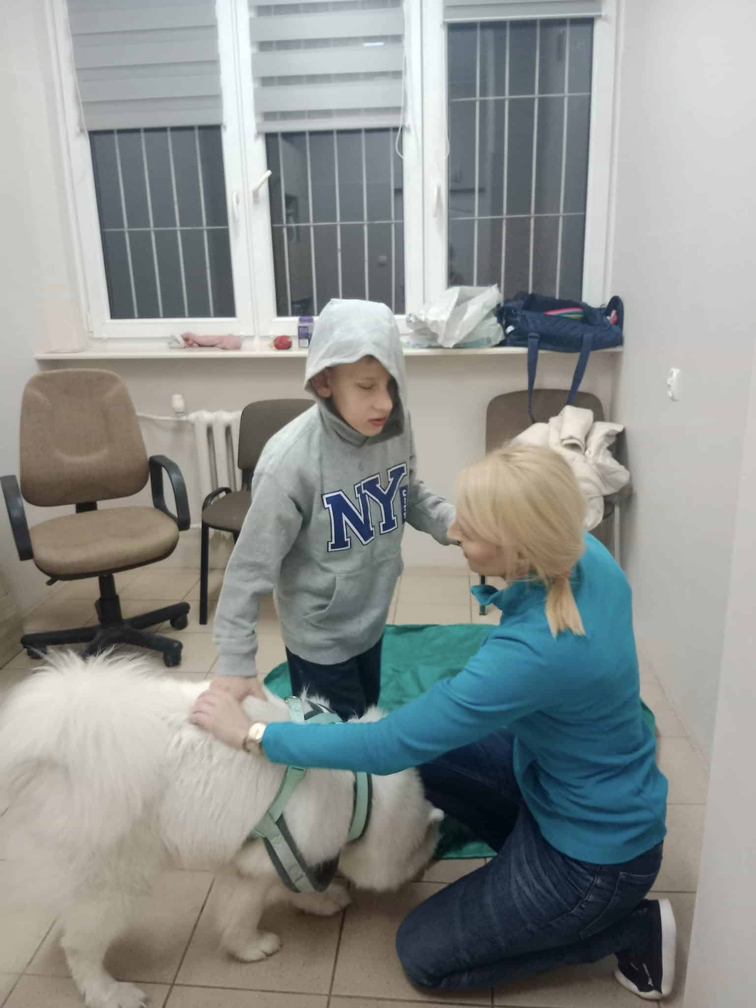 Kobieta klęczy obok młodego chłopca w bluzie z kapturem i białego psa w uprzęży w pokoju z krzesłami i oknem.