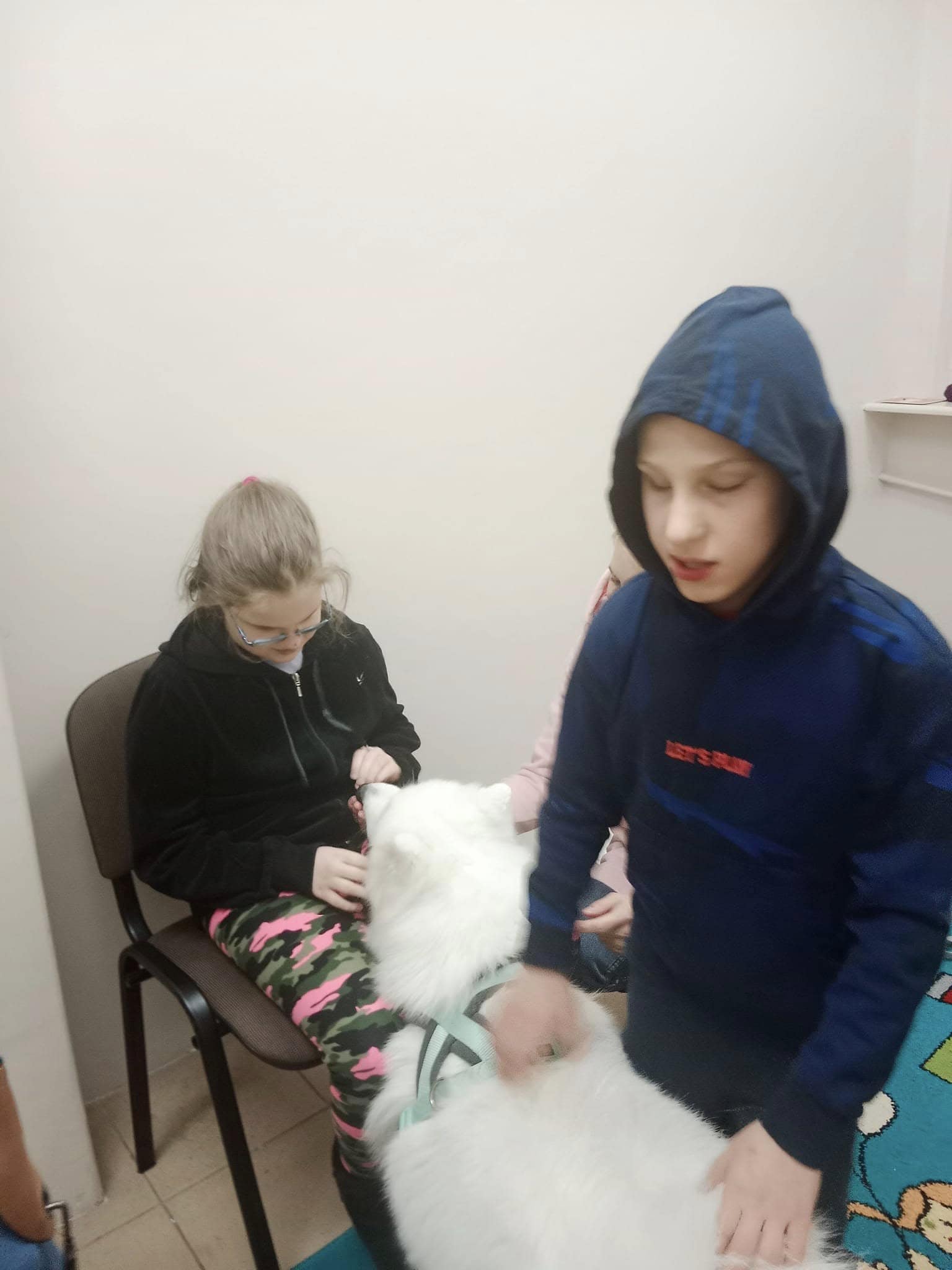 Dwoje dzieci jest w pomieszczeniu z dużym białym psem; jedno dziecko siedzi i głaszcze psa, podczas gdy drugie stoi w pobliżu.