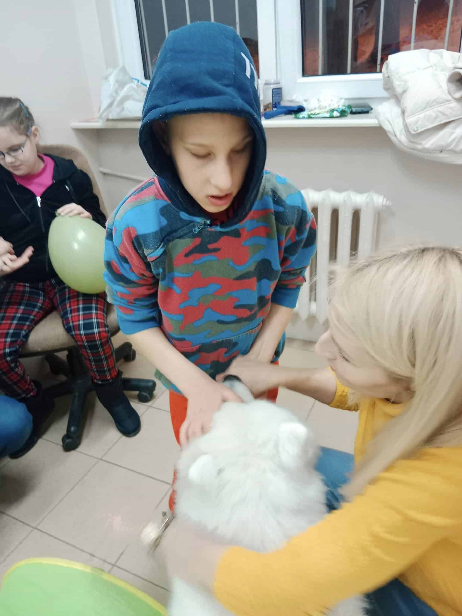 Chłopiec w niebiesko-czerwonej bluzie stoi obok kobiety głaszczącej białego psa. Inne dziecko siedzi w pobliżu, trzymając zielony balon.