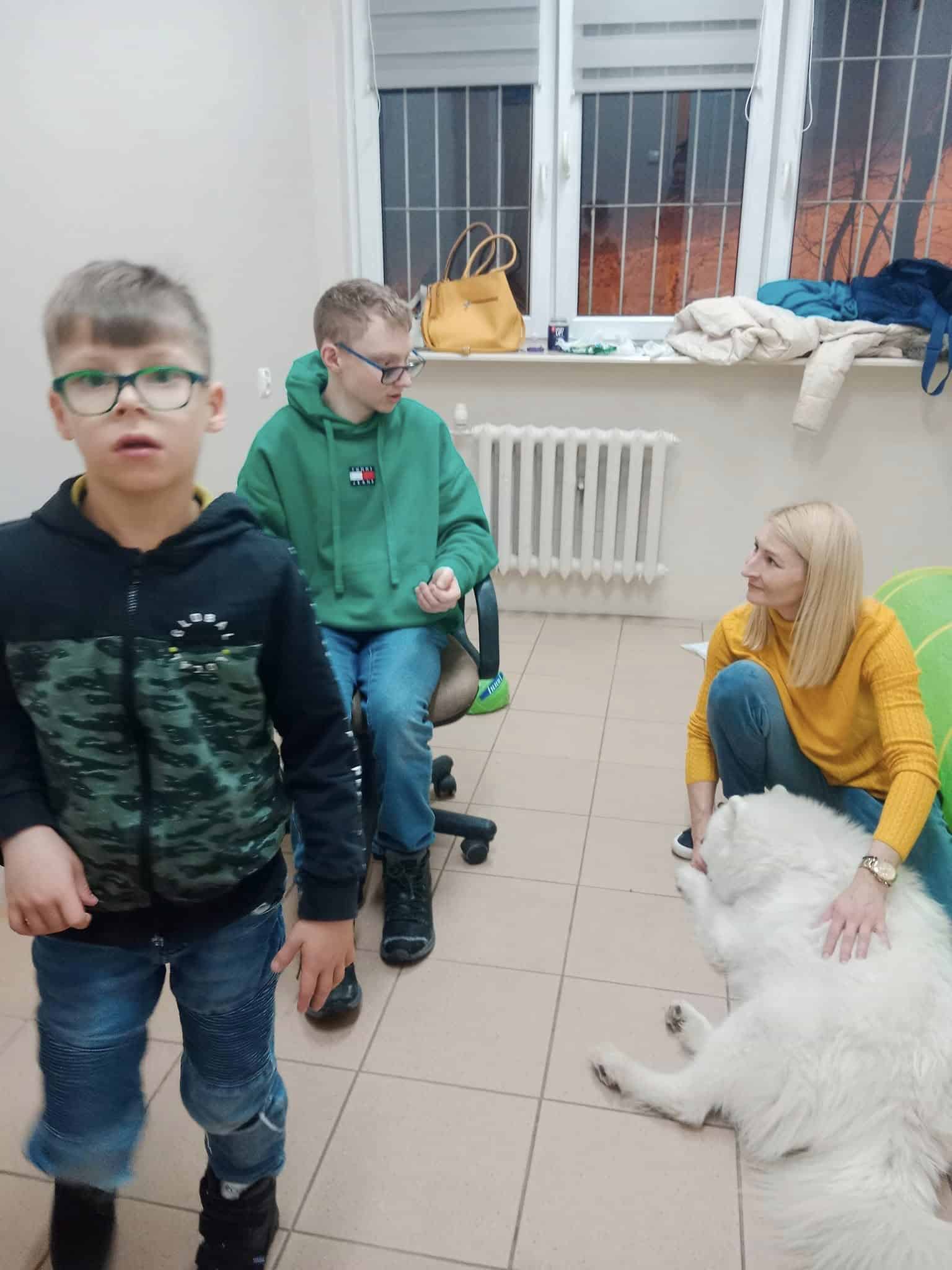 Trzy osoby i duży biały pies znajdują się w pokoju; kobieta siedzi na podłodze i głaszcze psa, podczas gdy dwóch chłopców jest w pobliżu, jeden siedzi, a drugi stoi.