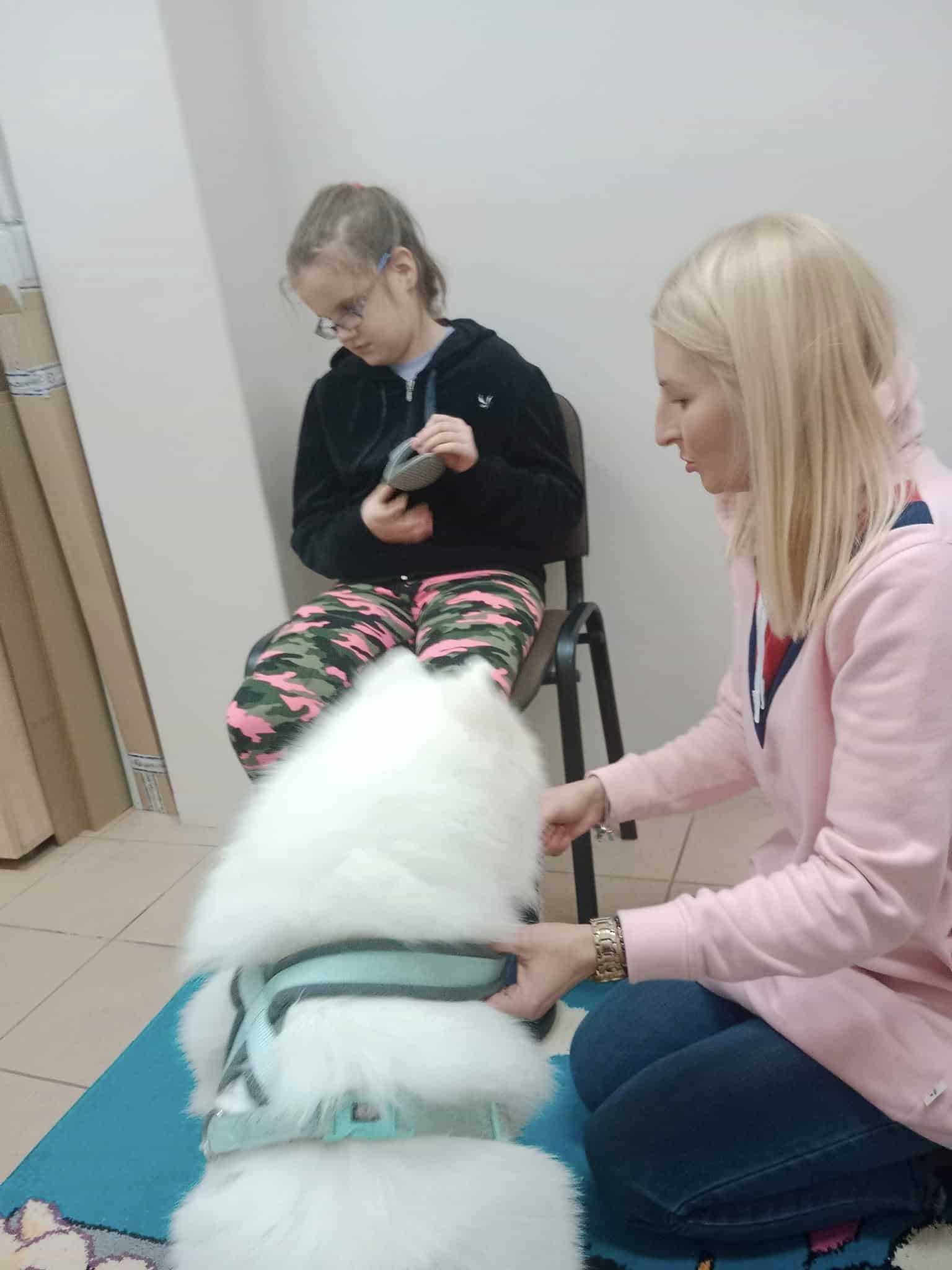 Kobieta klęczy obok puszystego białego psa w uprzęży, podczas gdy dziewczyna w okularach siedzi na krześle trzymając małe urządzenie.