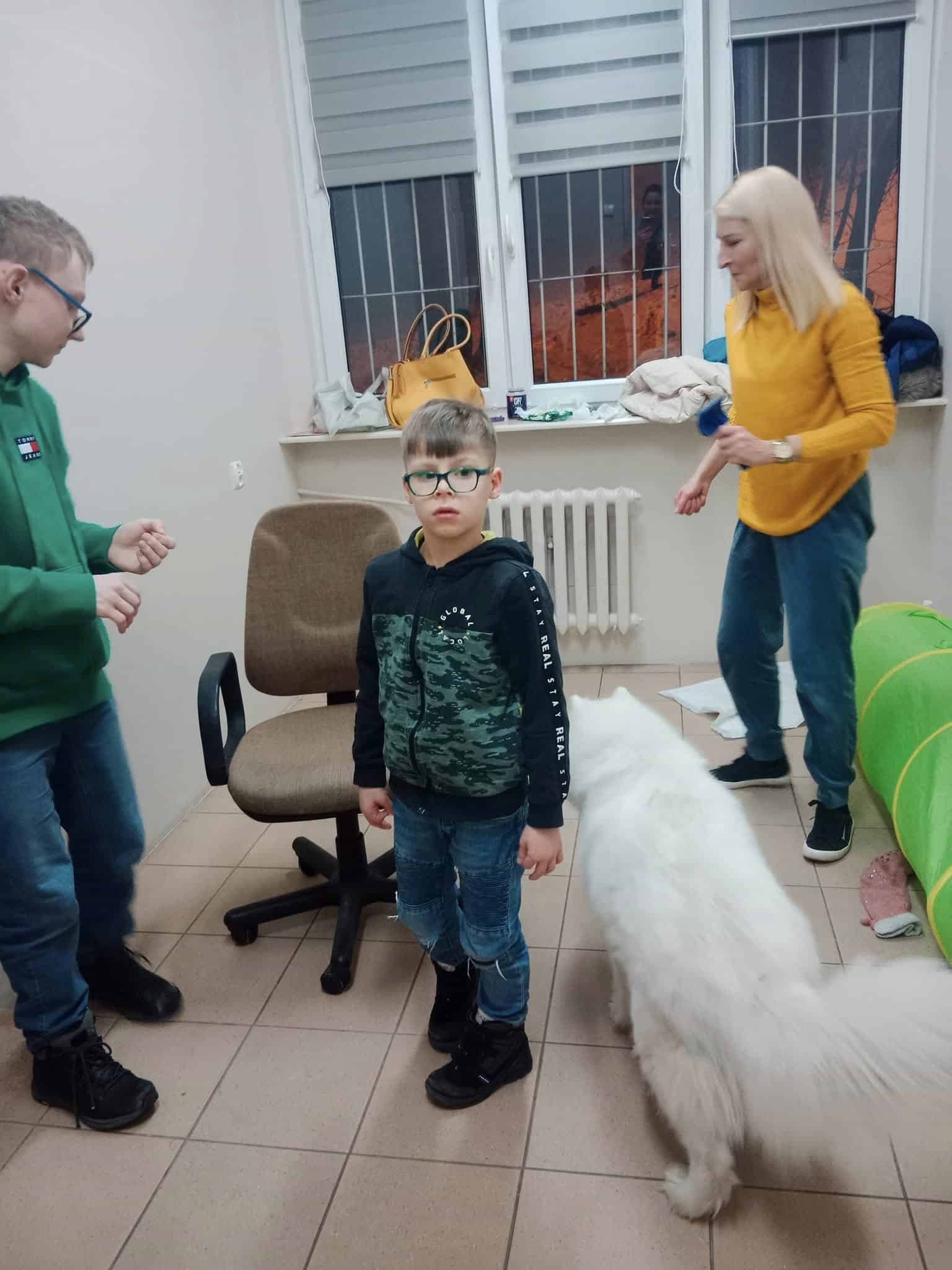 Chłopiec w okularach stoi w pokoju z fotelem, dwiema innymi osobami i dużym białym psem. Pokój ma podłogę wyłożoną kafelkami i okno z żaluzjami.