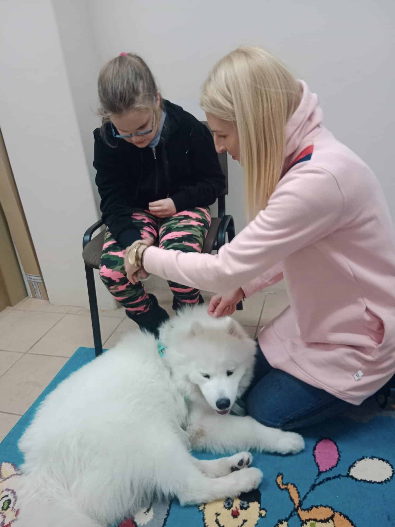 Kobieta i młoda dziewczyna wchodzą w interakcję z puszystym białym psem leżącym na kolorowej macie w pokoju z podłogą wyłożoną kafelkami.