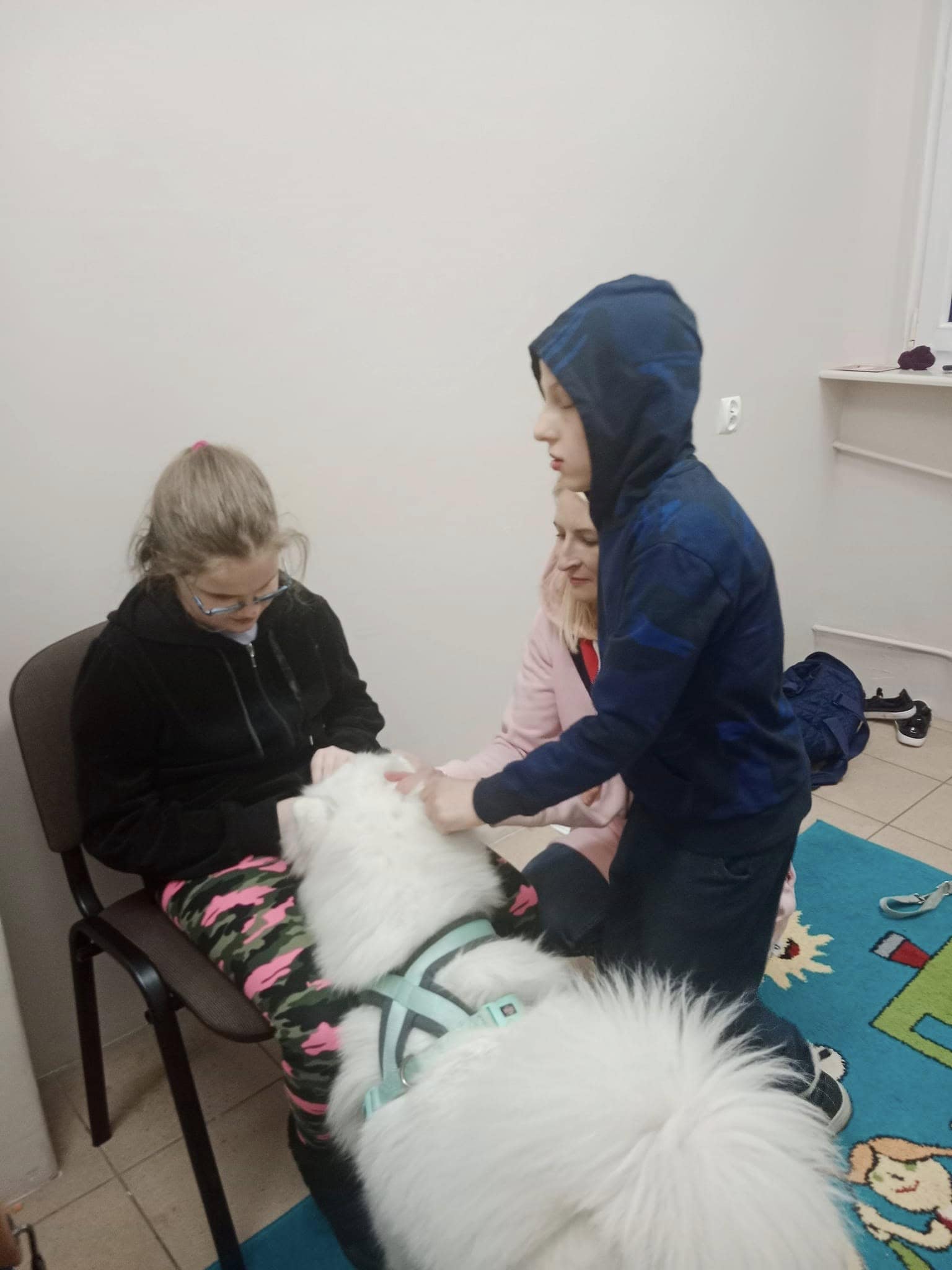 Dziewczyna siedzi na krześle, podczas gdy chłopiec głaszcze białego puszystego psa w uprzęży. W pobliżu klęczy inna kobieta. Znajdują się w małym, prostym pokoju z zabawkami na podłodze.