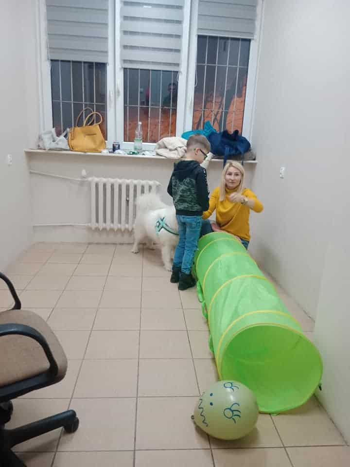 Kobieta i dziecko wchodzą w interakcję w pobliżu zielonego tunelu do zabawy w pokoju z białym psem, balonami, krzesłem i oknem z kratami.