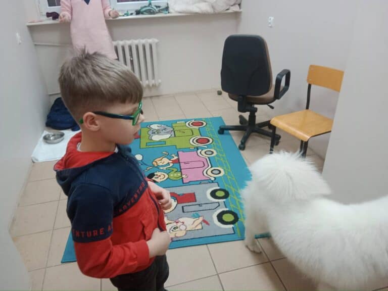 Chłopiec w okularach stoi obok białego puszystego psa na kolorowym dywanie w małym pokoju z krzesłami i oknem.