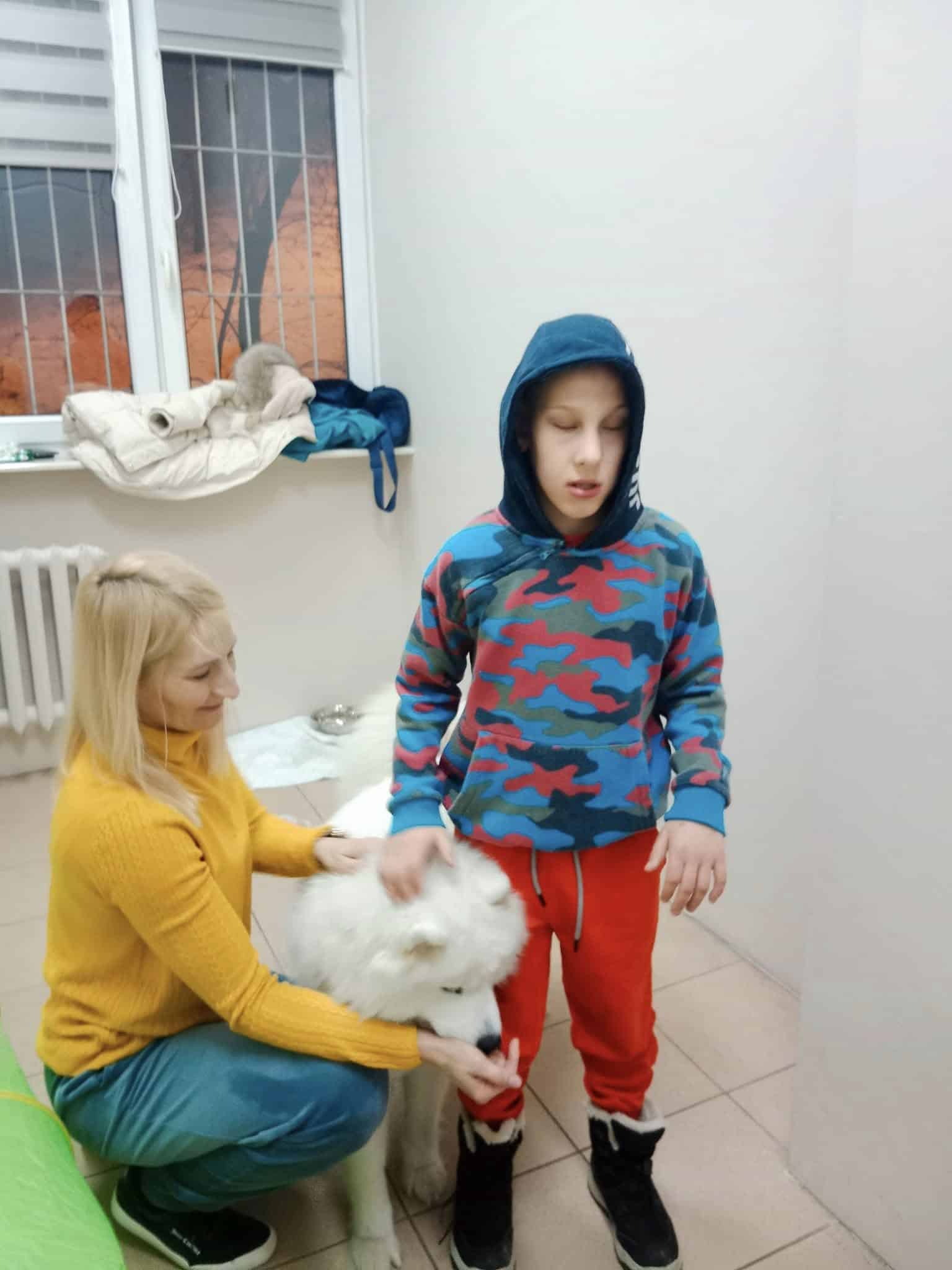 Kobieta klęczy obok białego psa i chłopca w niebiesko-czerwonej bluzie, wewnątrz pokoju z białymi ścianami i oknem z kratami.