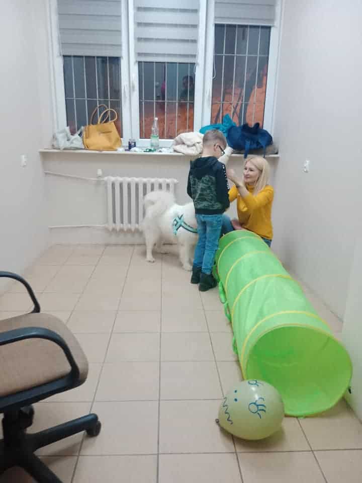 Kobieta klęczy obok małego chłopca i białego psa w małym pokoju z zielonym tunelem do zabawy i balonem na podłodze.