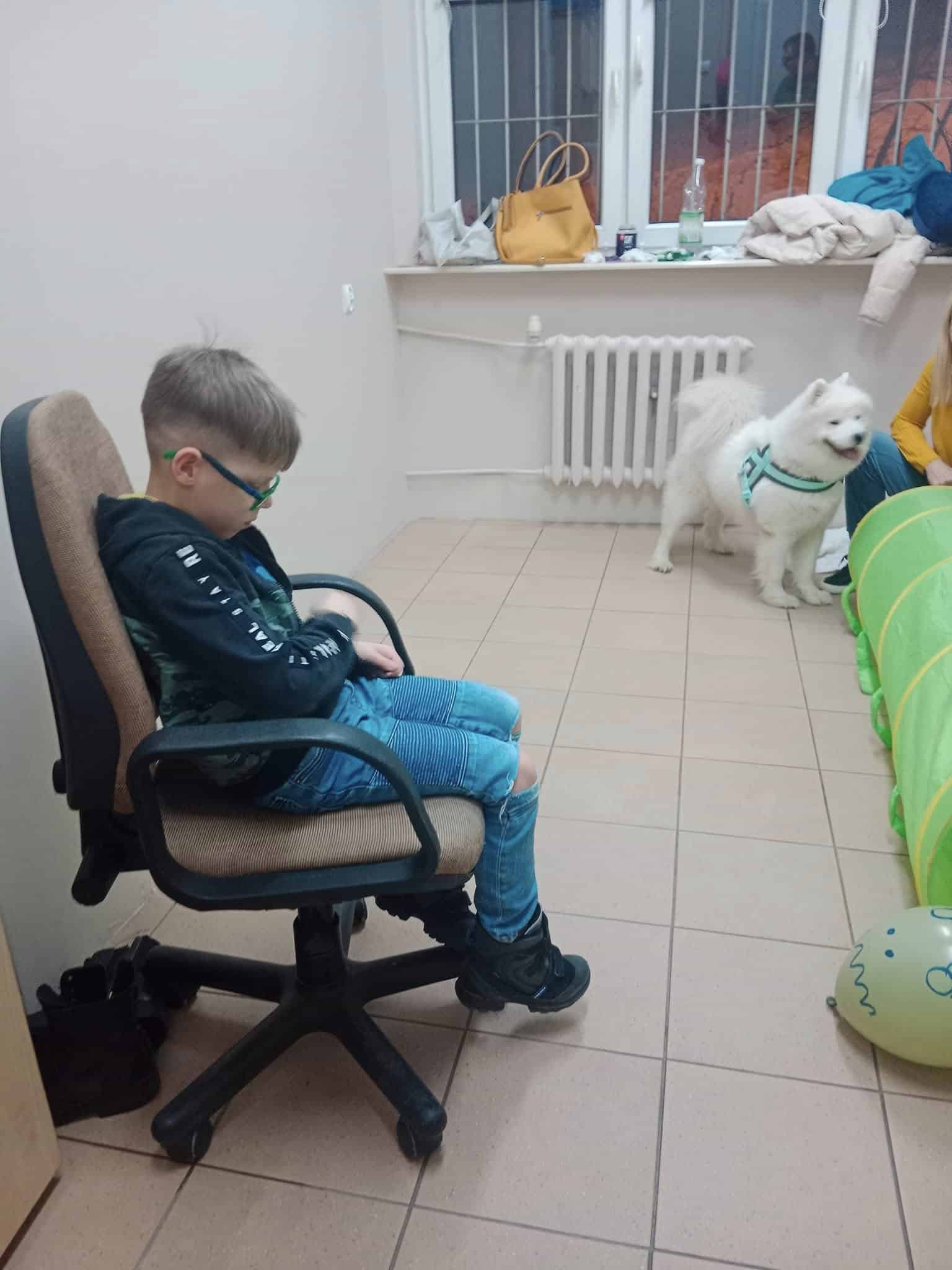 Chłopiec w okularach siedzi na krześle biurowym, patrząc w dół. Puszysty biały pies stoi w pobliżu, obok kobiety częściowo widocznej przez zabawkę tunelową i balony.