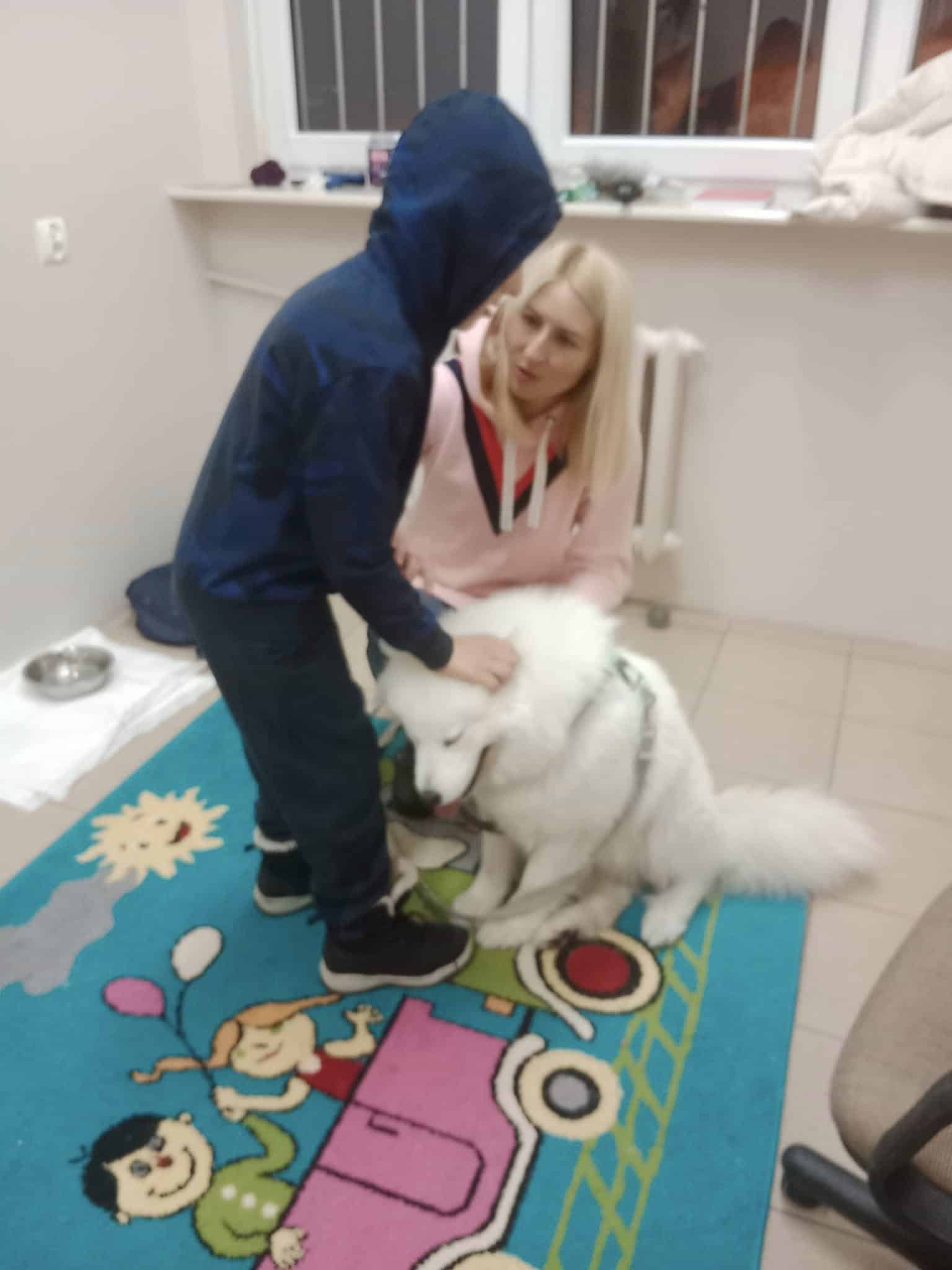 Chłopiec w niebieskiej bluzie z kapturem głaszcze białego psa, podczas gdy kobieta o blond włosach klęczy obok nich na kolorowym dywanie w pokoju.