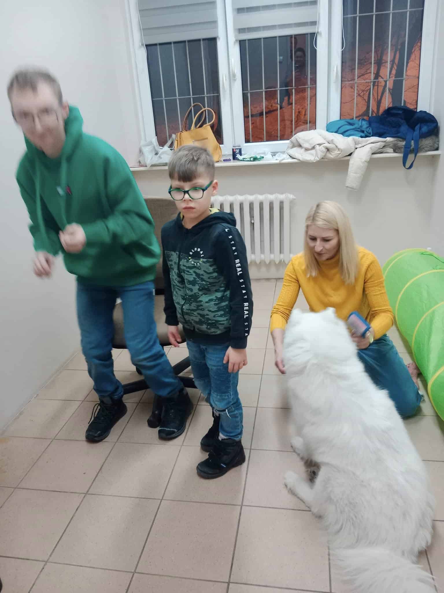 Dwoje dorosłych i dziecko stoją w pokoju z dużym białym psem; jeden dorosły klęczy, trzymając telefon, a dziecko patrzy w kamerę.