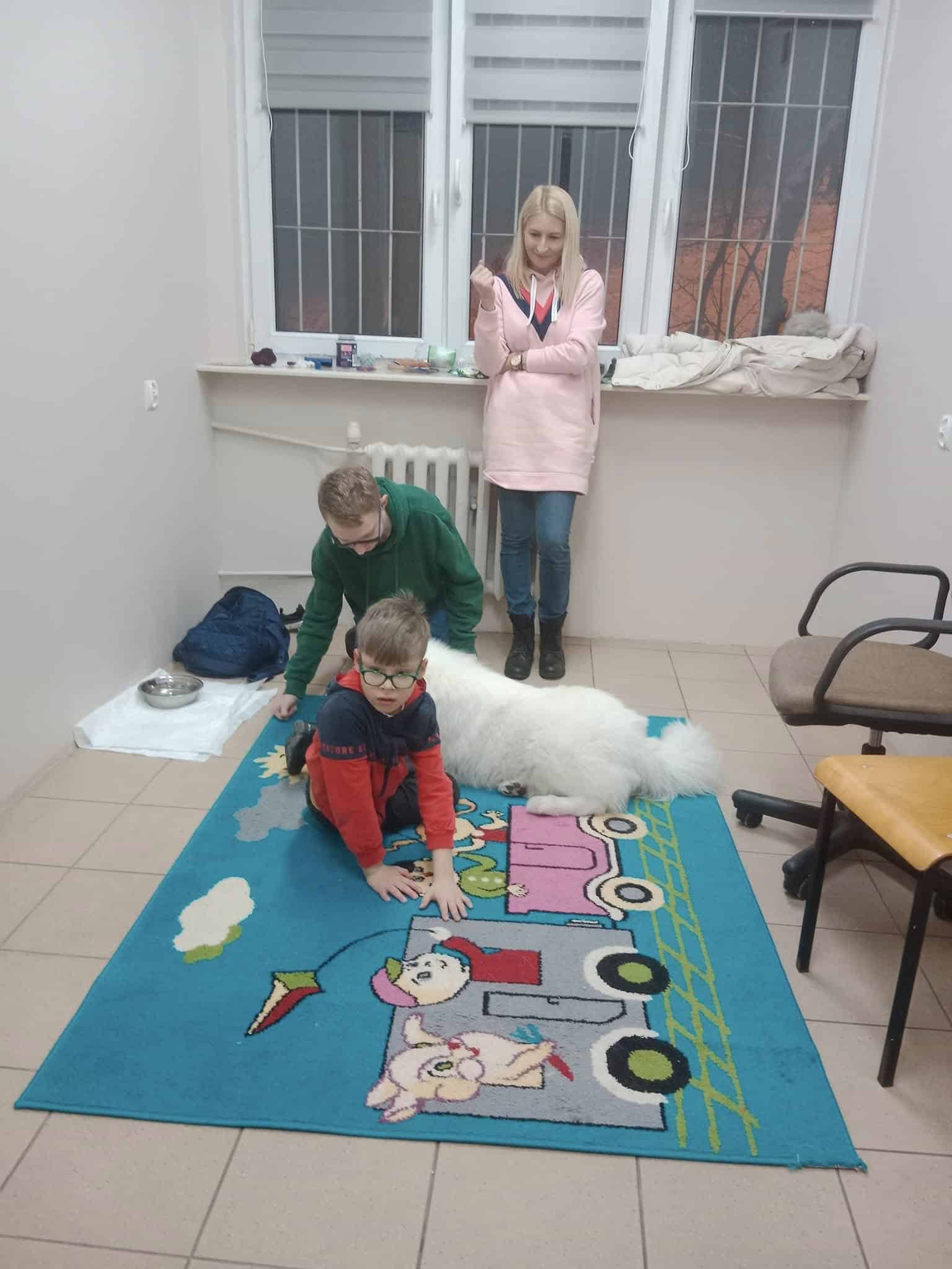 Dwoje dzieci bawi się na kolorowym dywanie z białym psem, podczas gdy kobieta stoi w pobliżu w słabo umeblowanym pokoju z zakratowanymi oknami.