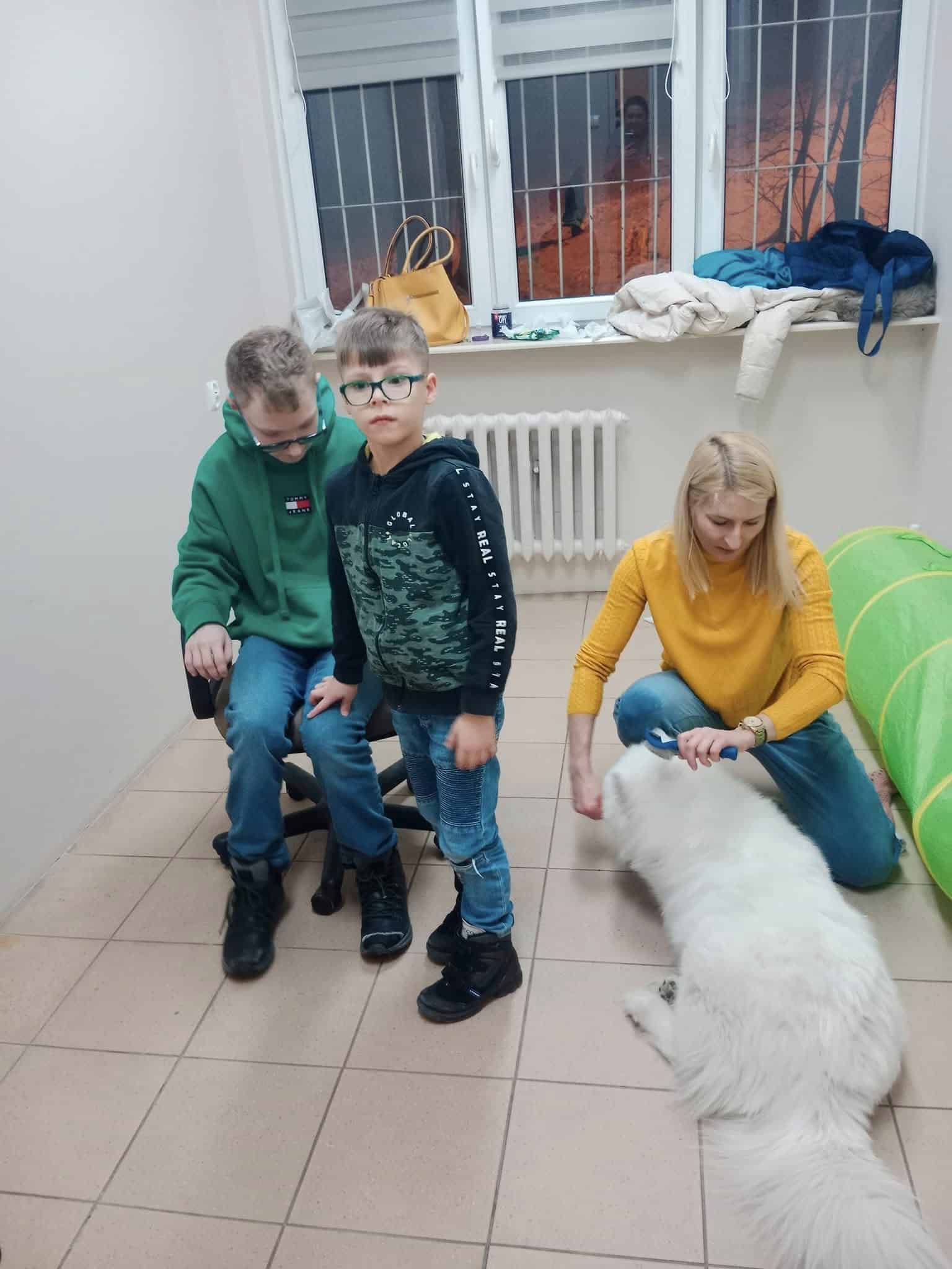 Dwóch chłopców i kobieta znajdują się w pokoju, w którym na podłodze leży duży biały pies. Kobieta głaszcze psa, podczas gdy chłopcy stoją w pobliżu. Płaszcze i torby leżą na parapecie.