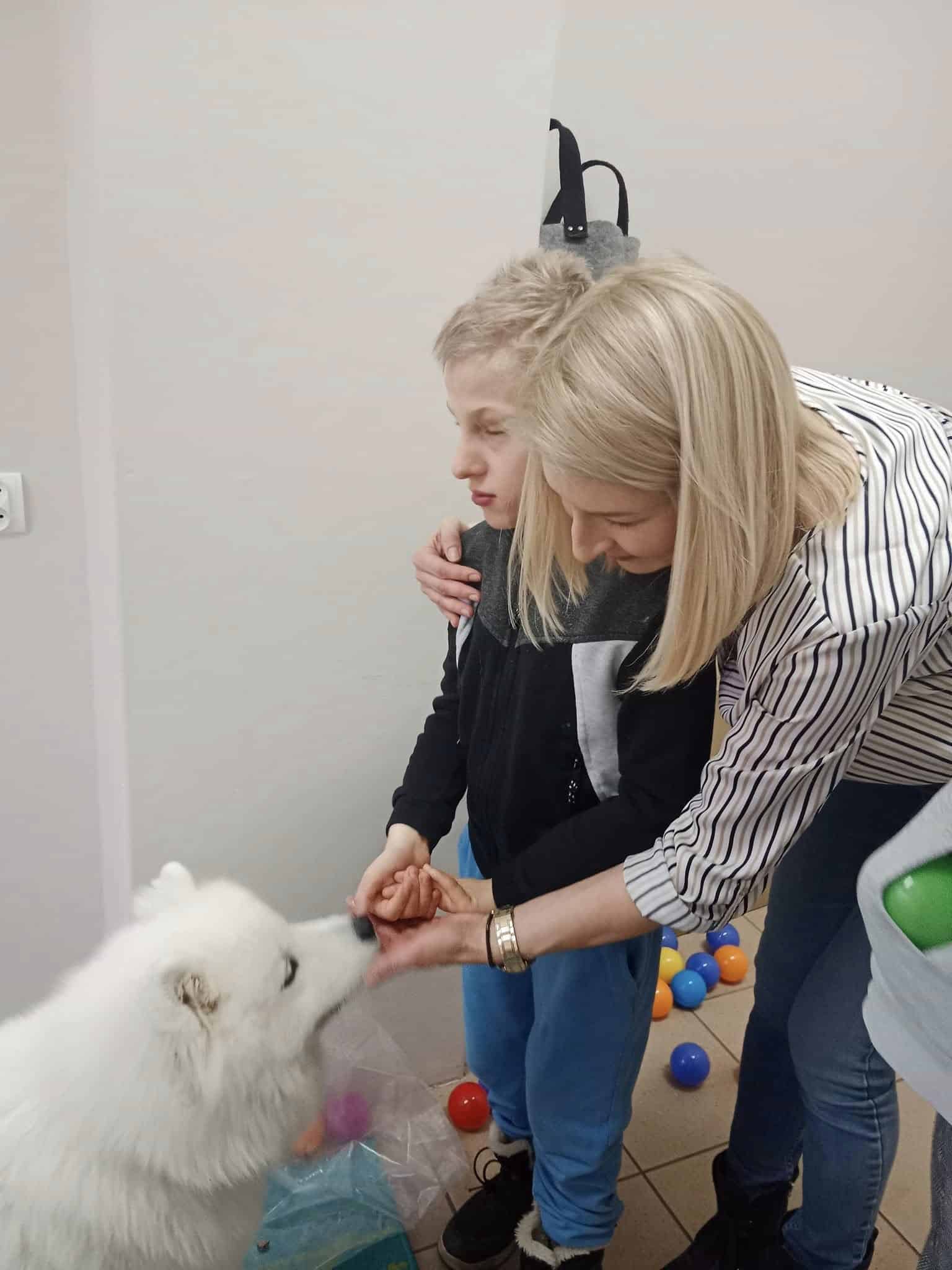 Kobieta pomaga chłopcu nakarmić białego puszystego psa w domu, z kolorowymi plastikowymi piłkami rozrzuconymi na podłodze w tle.