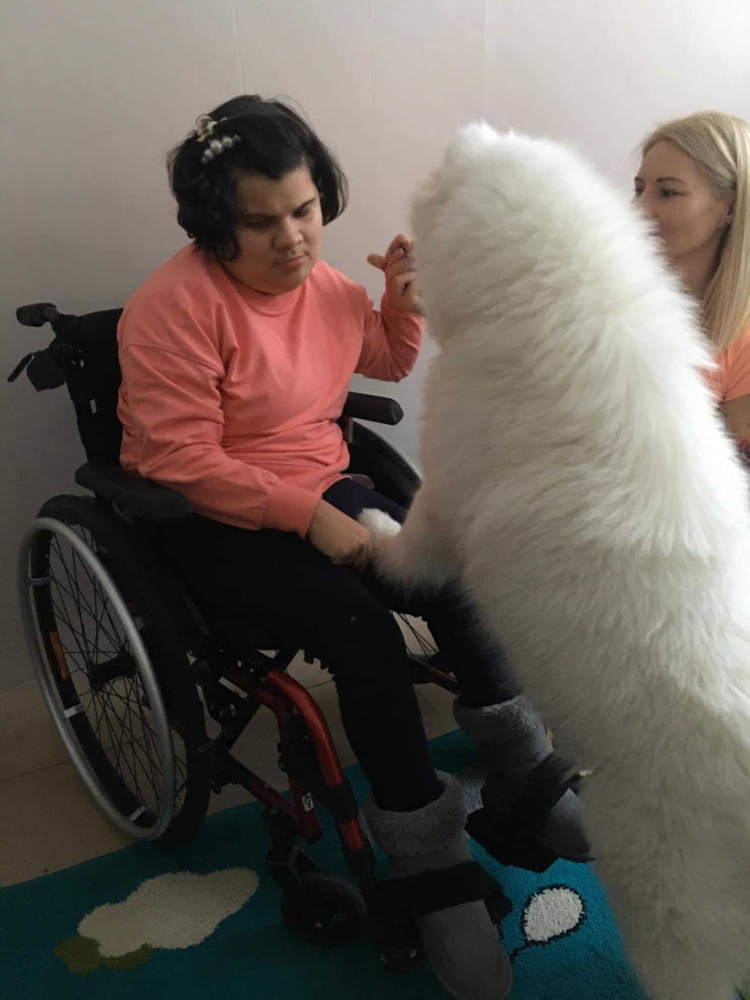Młoda kobieta na wózku inwalidzkim głaszcze dużego, puszystego białego psa, a obok niej jest inna kobieta.
