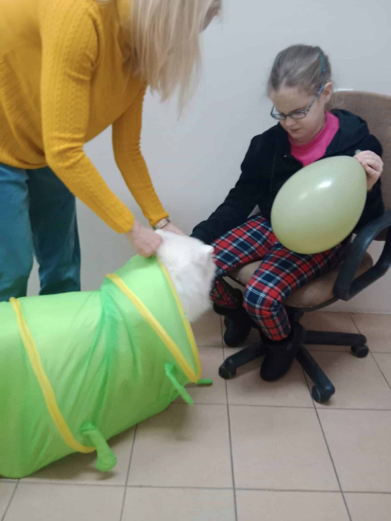 Dorosły pomaga psu przejść przez zielony tunel do zabawy, podczas gdy dziewczynka w okularach trzymająca żółty balon siedzi na krześle.