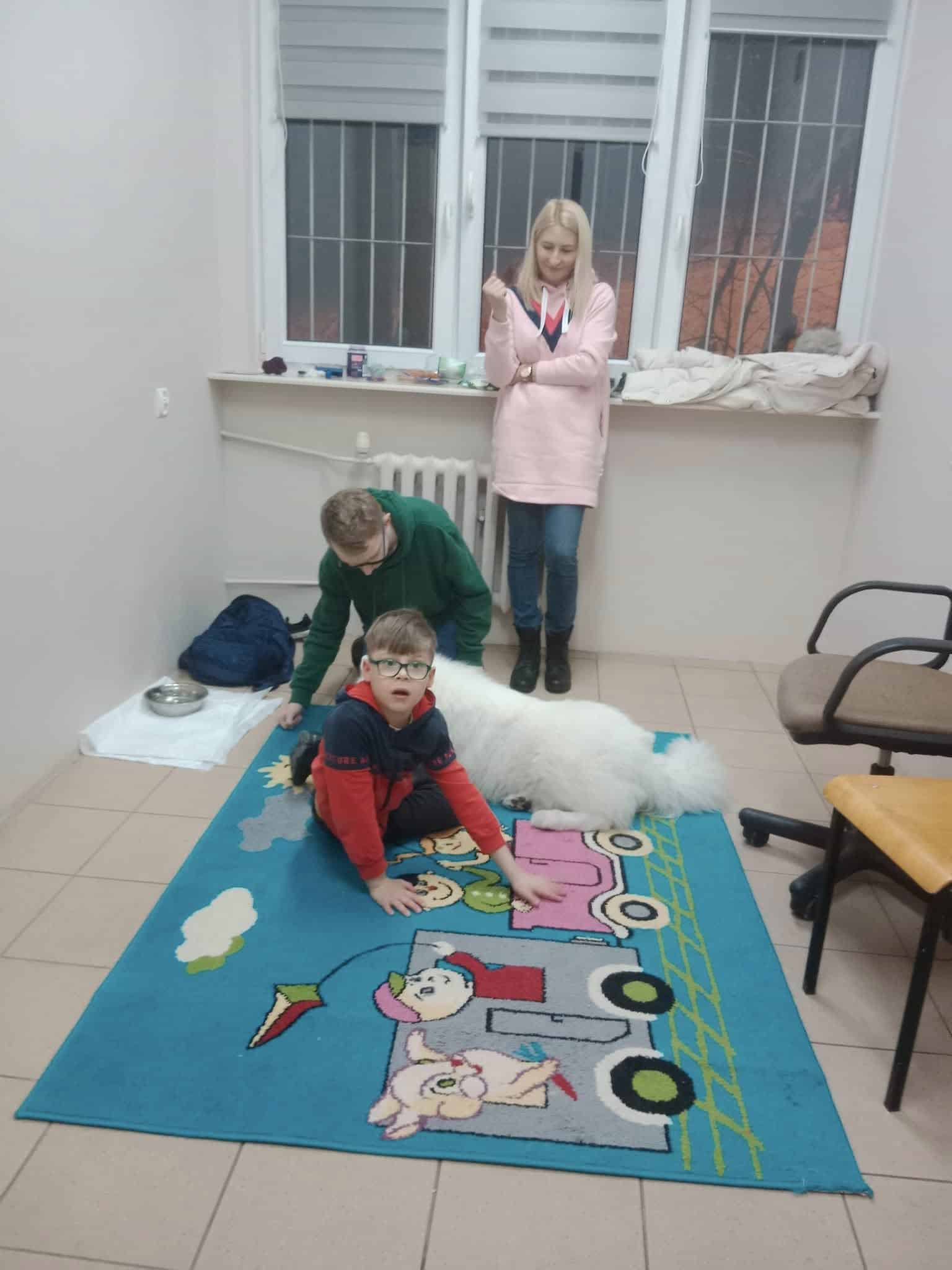 Dwoje dzieci i kobieta znajdują się w małym pokoju z podłogą wyłożoną kafelkami. Jedno dziecko siedzi na kolorowym dywanie z motywem pociągu, podczas gdy drugie pochyla się nad puszystym białym psem. Kobieta stoi w pobliżu.