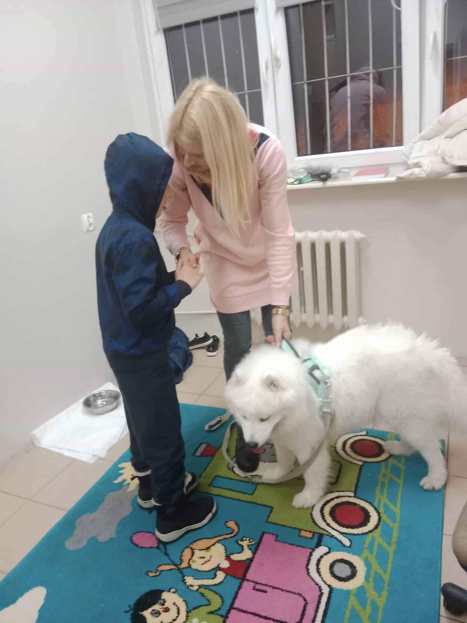 Kobieta i dziecko stoją na kolorowym dywanie z białym puszystym psem w uprzęży w pokoju z zakratowanymi oknami i kaloryferem.