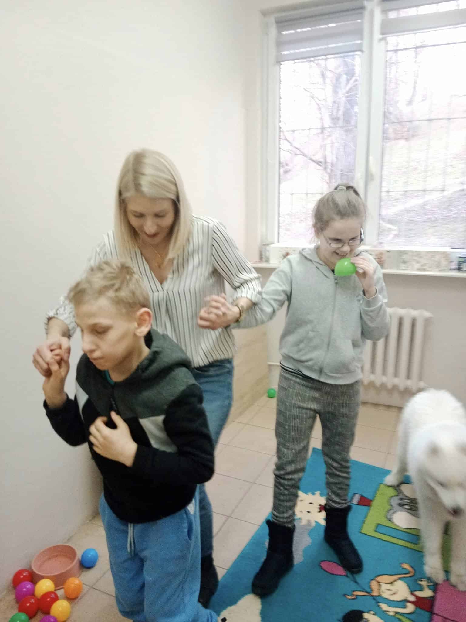 Dorosły trzyma się za ręce z dwójką dzieci spacerujących po pokoju z kolorowymi piłkami i zabawnym dywanem; w pobliżu stoi biały pies.