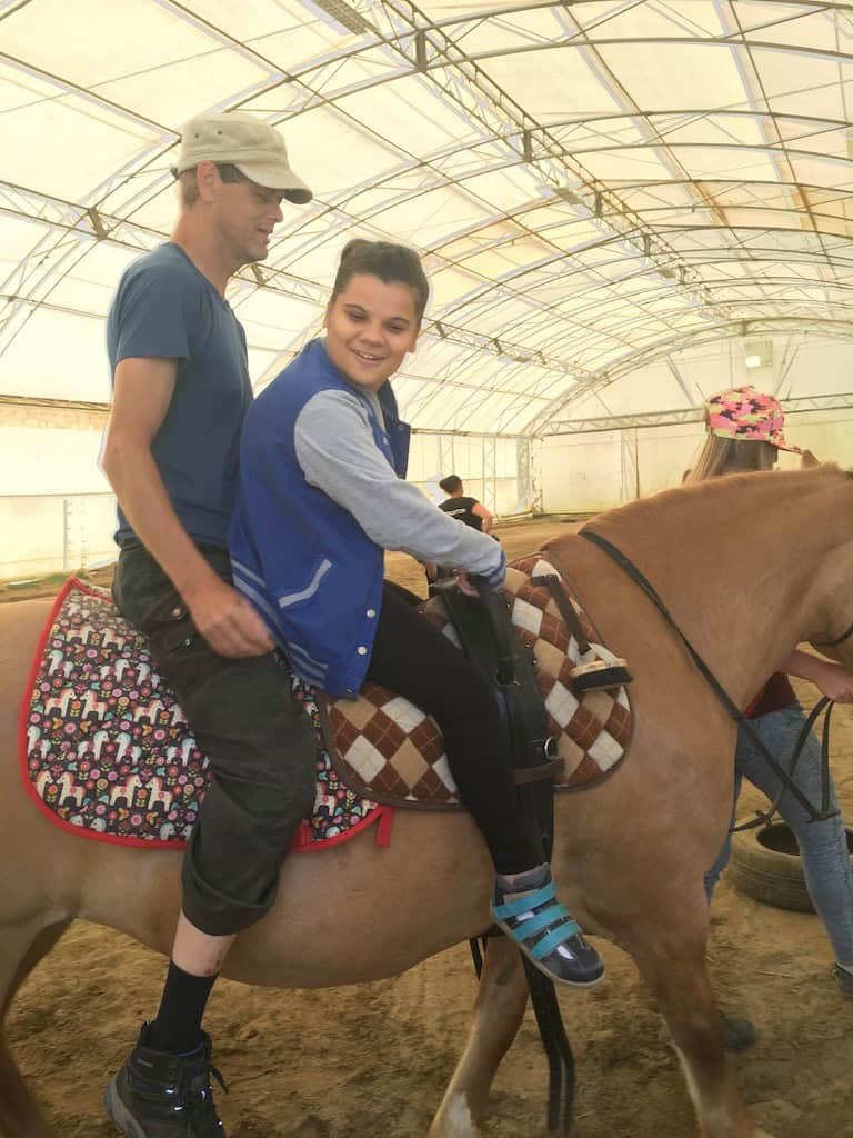 Mężczyzna i uśmiechnięta młoda osoba siedzą razem na jasnobrązowym koniu na zadaszonej ujeżdżalni. Inna osoba stoi w pobliżu, częściowo poza kadrem.