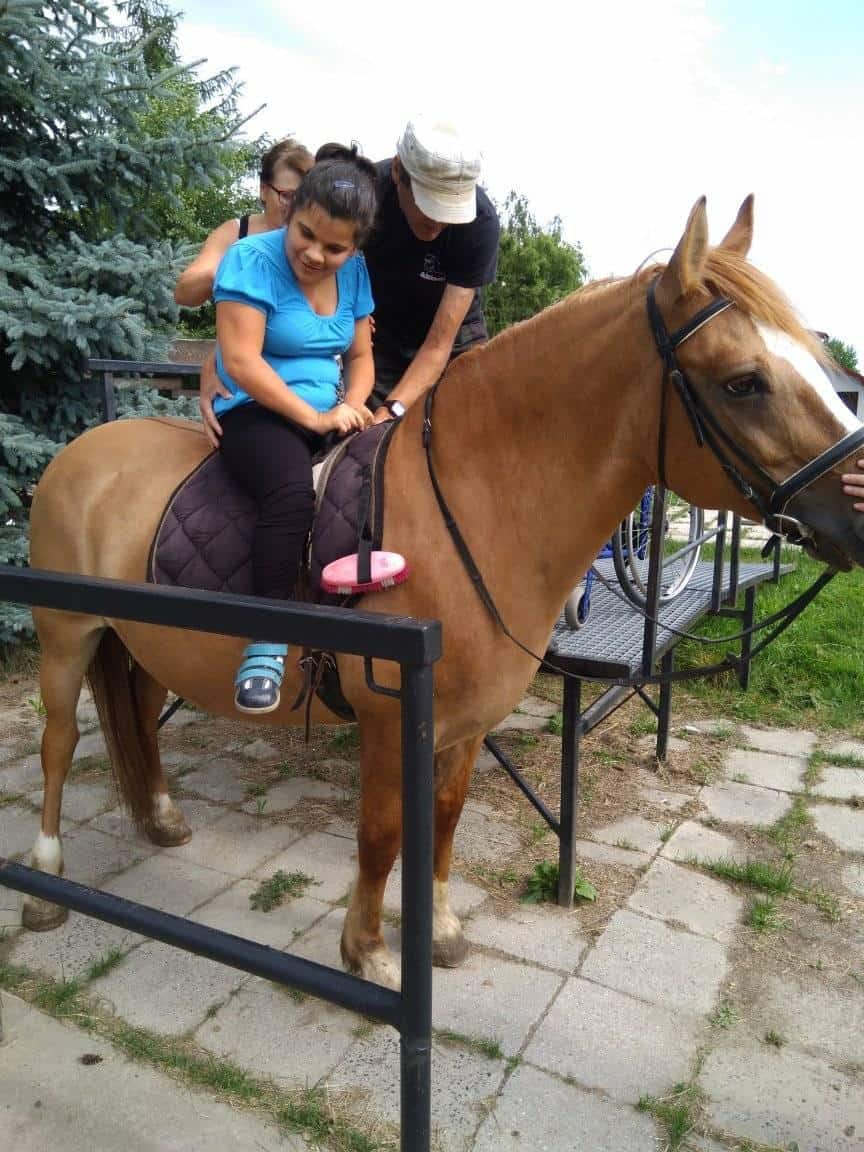 Młoda kobieta siedzi na osiodłanym koniu z pomocą dwóch dorosłych osób na utwardzonym terenie.