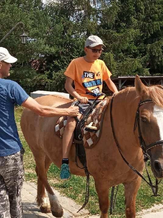 Chłopiec w okularach przeciwsłonecznych i pomarańczowej koszulce siedzi na brązowym koniu, podczas gdy dorosły w niebieskiej koszuli i czapce trzyma uzdę konia na zewnątrz.