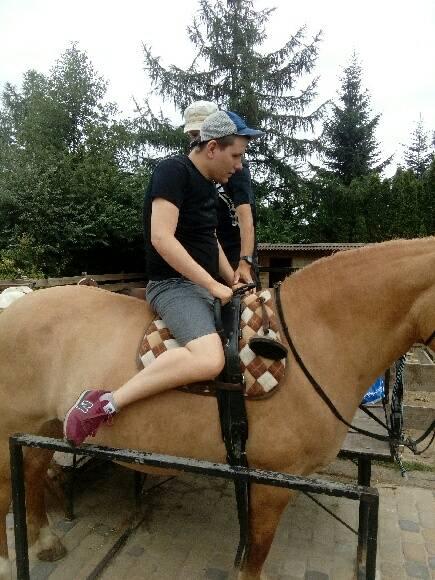 Osoba ubrana w czarną koszulę, szare szorty i czapkę siedzi bokiem na opalonym koniu na zewnątrz w pobliżu drzew.