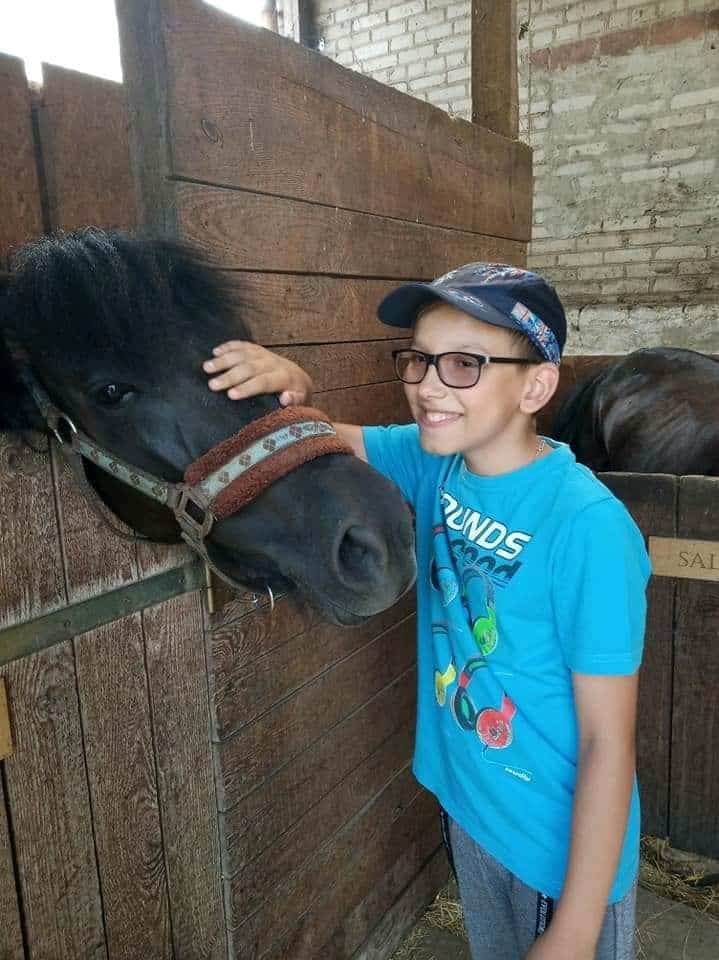 Chłopiec w okularach i niebieskiej koszulce uśmiecha się, głaszcząc czarnego konia w drewnianej stajni.