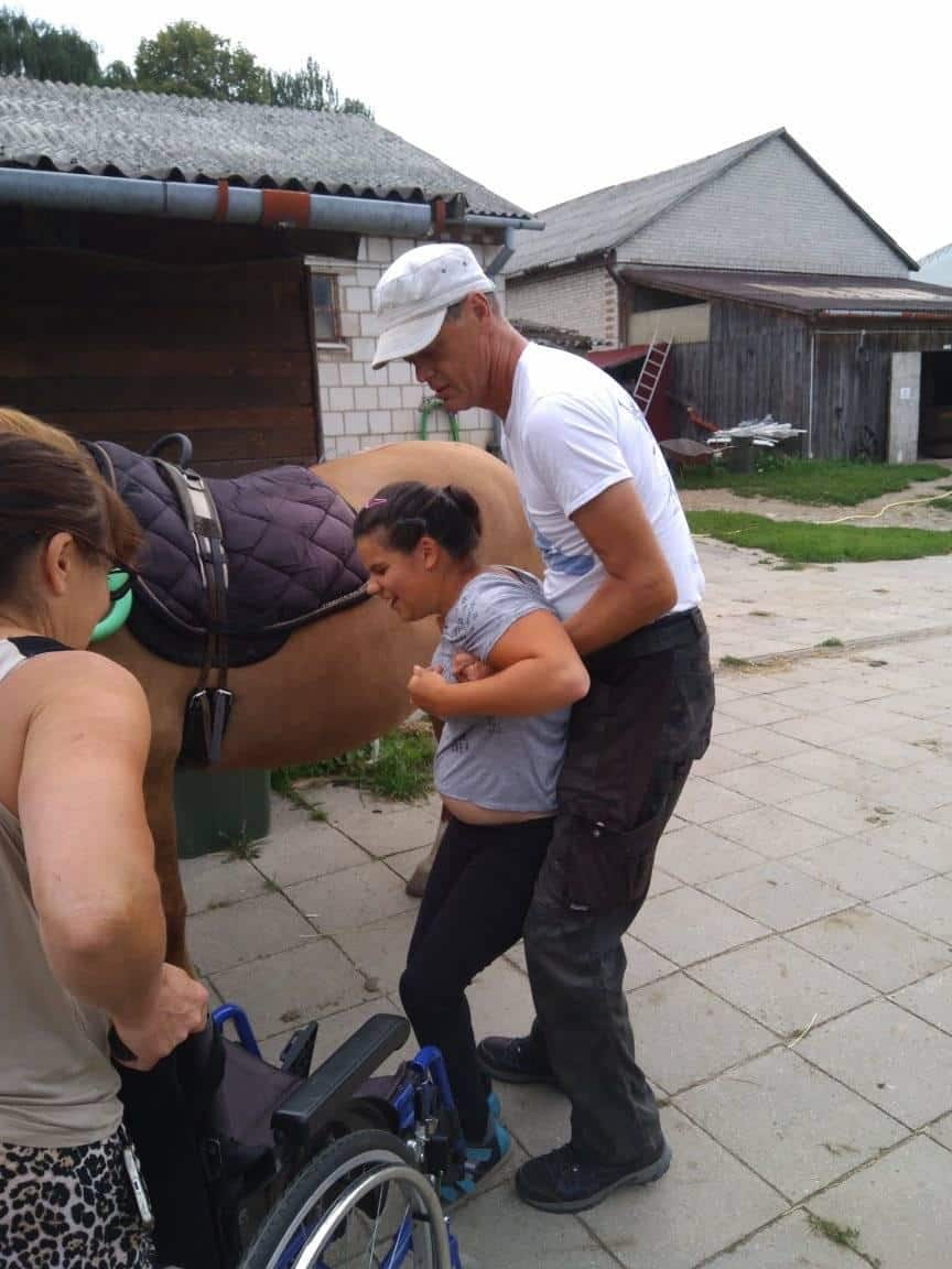 Mężczyzna wspiera kobietę wstającą z wózka inwalidzkiego obok osiodłanego konia, podczas gdy inna kobieta pomaga, na zewnątrz w pobliżu stajni.