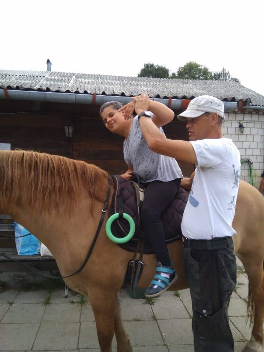 Osoba pomaga kobiecie w ćwiczeniach rozciągających, podczas gdy ona siedzi na brązowym koniu na zewnątrz w pobliżu budynku.