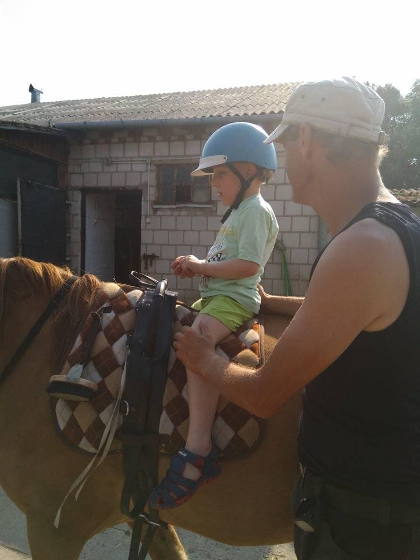 Dorosły pomaga małemu dziecku w kasku usiąść na koniu na zewnątrz w pobliżu budynku.