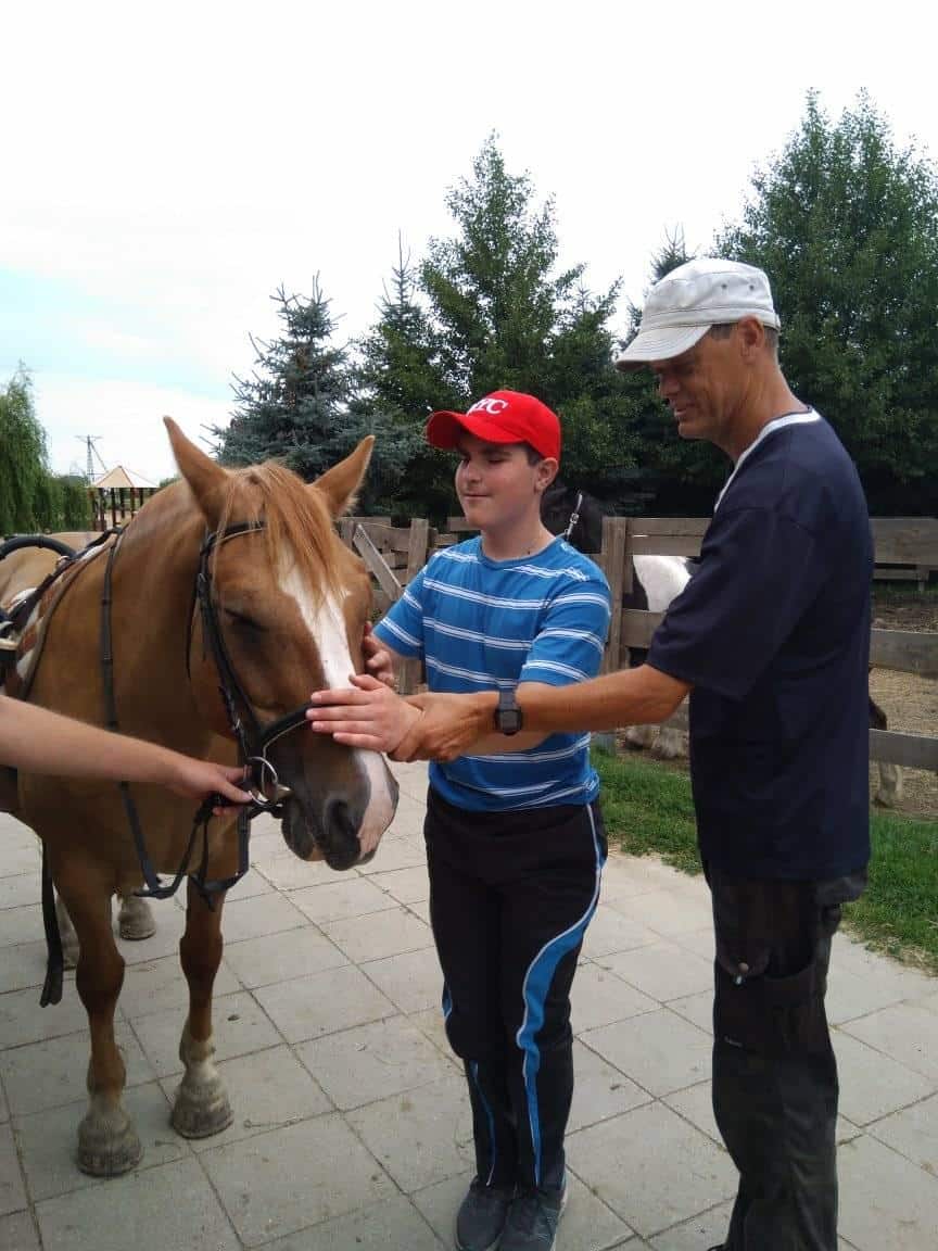 Chłopiec w czerwonej czapce i mężczyzna w białym kapeluszu stoją obok brązowego konia, głaszcząc go po głowie na zewnątrz w pobliżu ogrodzenia i kilku drzew.