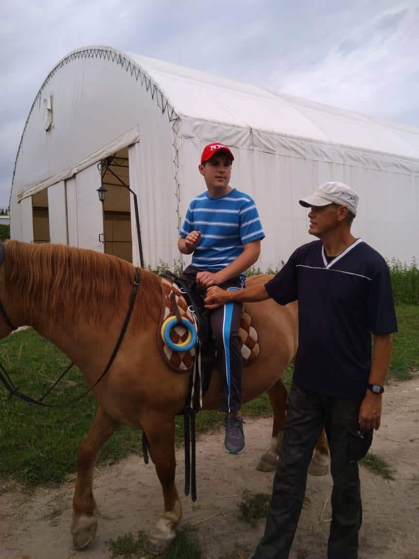 Chłopiec w pasiastej koszuli i czerwonej czapce siedzi na brązowym koniu, podczas gdy mężczyzna w czapce trzyma konia w pobliżu białego budynku przypominającego namiot.