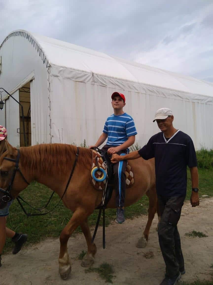 Chłopiec w pasiastej koszuli i czerwonej czapce jedzie na brązowym koniu, prowadzony przez mężczyznę w białym kapeluszu, w pobliżu dużego białego namiotu w pochmurny dzień.