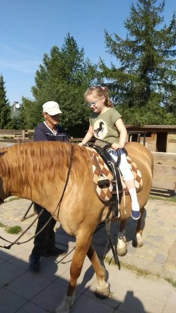 Dziecko siedzi na brązowym koniu, prowadzonym przez dorosłego mężczyznę na zewnątrz w słoneczny dzień, z drzewami i stajniami w tle.