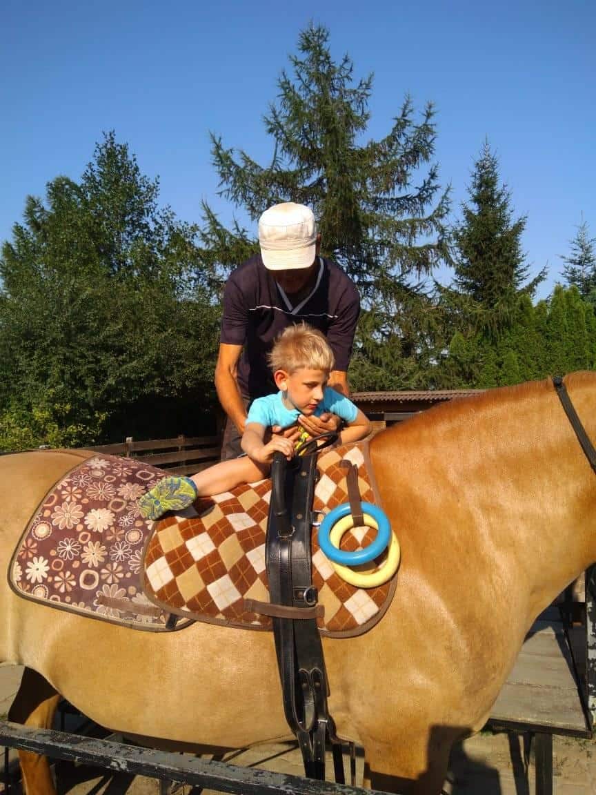 Dorosły pomaga młodemu chłopcu usiąść na osiodłanym koniu na świeżym powietrzu, z drzewami i czystym niebem w tle. Chłopiec trzyma kolorowe pierścionki.