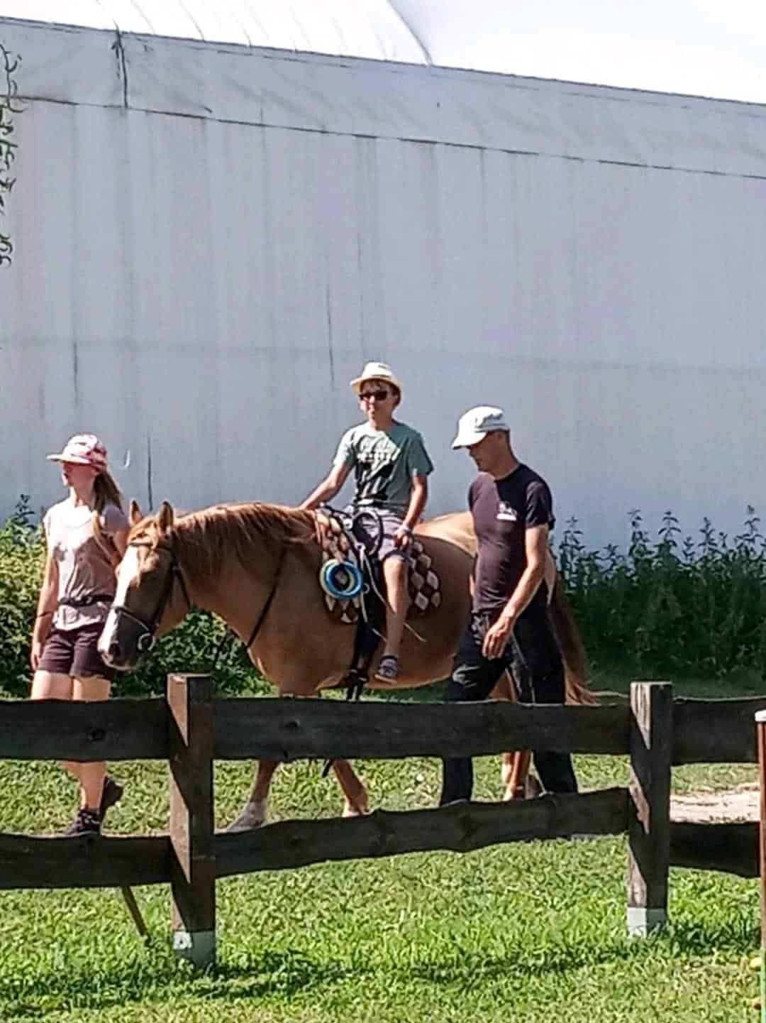 Osoba jedzie na jasnobrązowym koniu, podczas gdy dwoje dorosłych idzie obok, jeden prowadzi konia, w słoneczny dzień w pobliżu drewnianego ogrodzenia i białego budynku.
