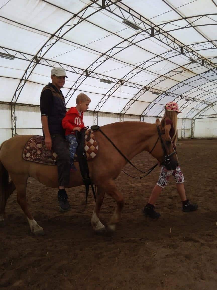 Dorosły i dziecko siedzą na koniu prowadzonym przez młodą dziewczynę na zadaszonej ujeżdżalni.